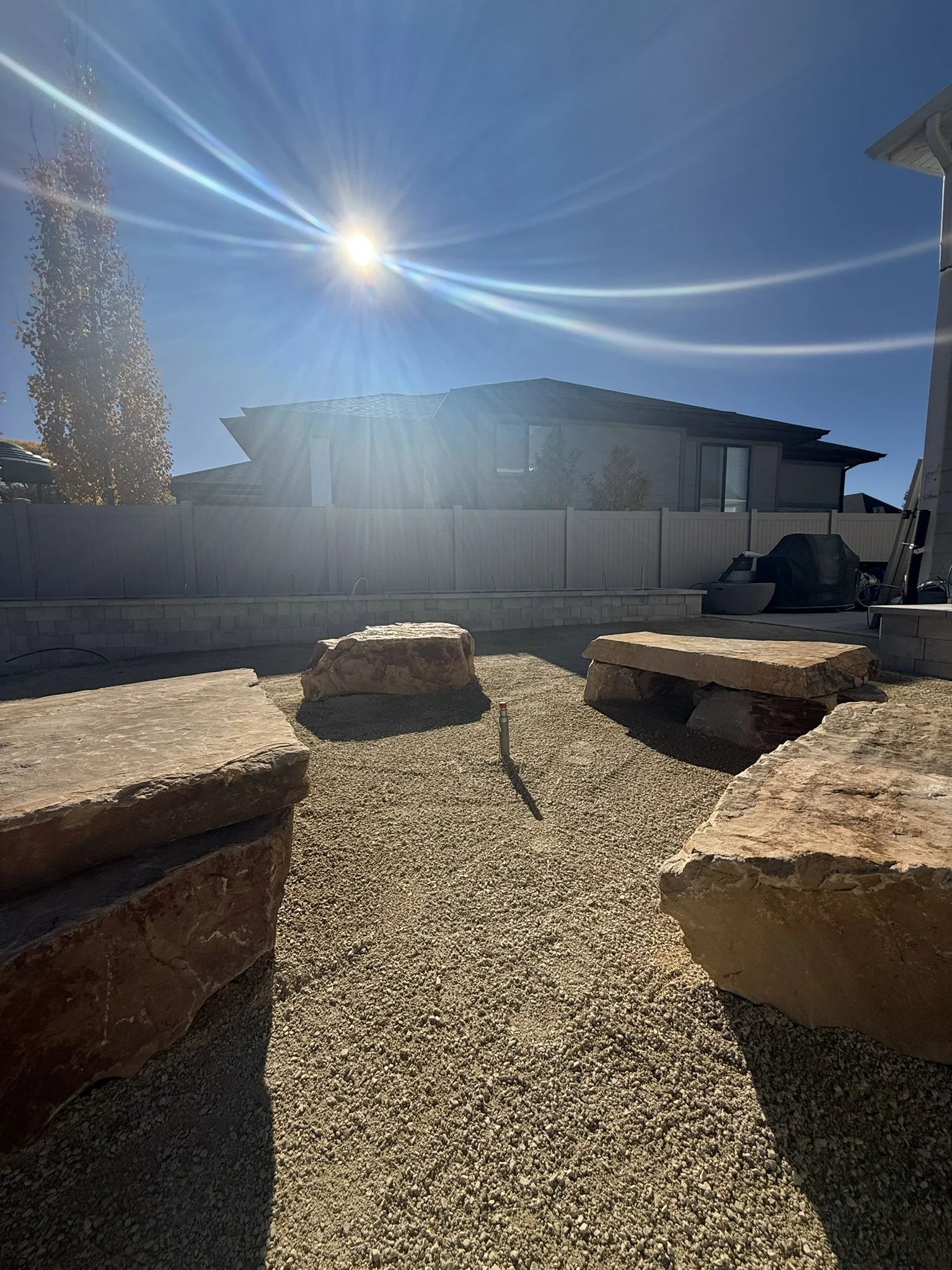 A backyard with a gravel ground, large rocks arranged as seating, a house in the background, and a bright sun in a clear blue sky creating lens flare.