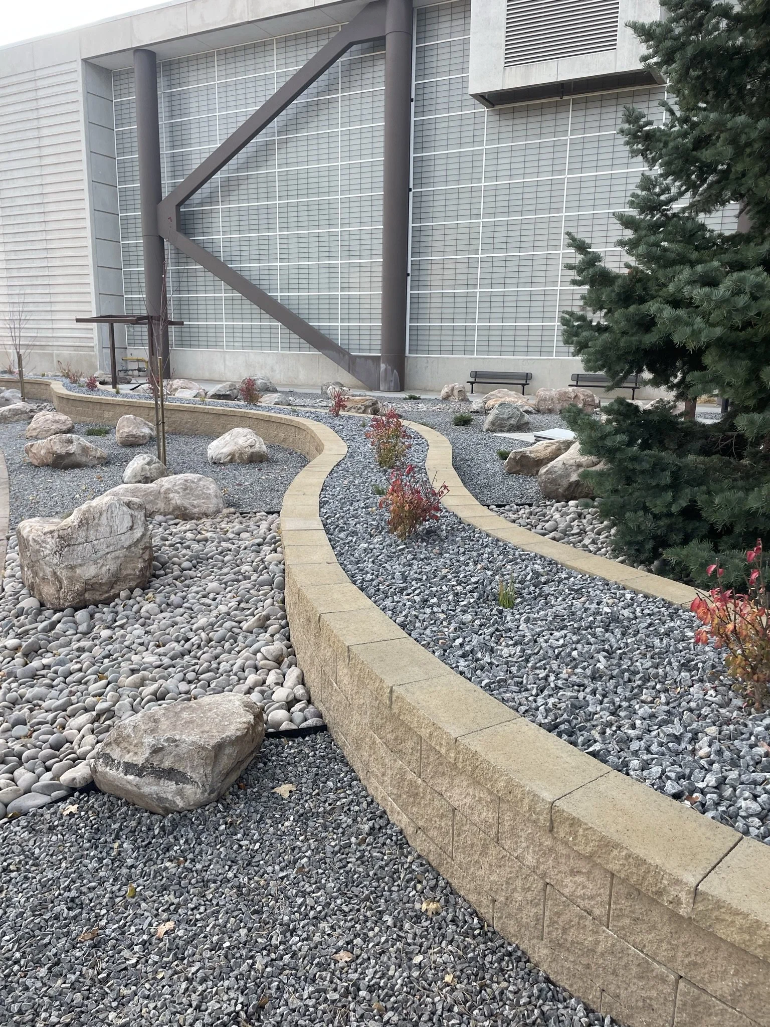 A landscaped outdoor area featuring a curved stone and gravel pathway bordered by a low beige brick wall, with large rocks and small plants along the path, in front of a modern building with large glass windows and metal beams, and a tall pine tree o