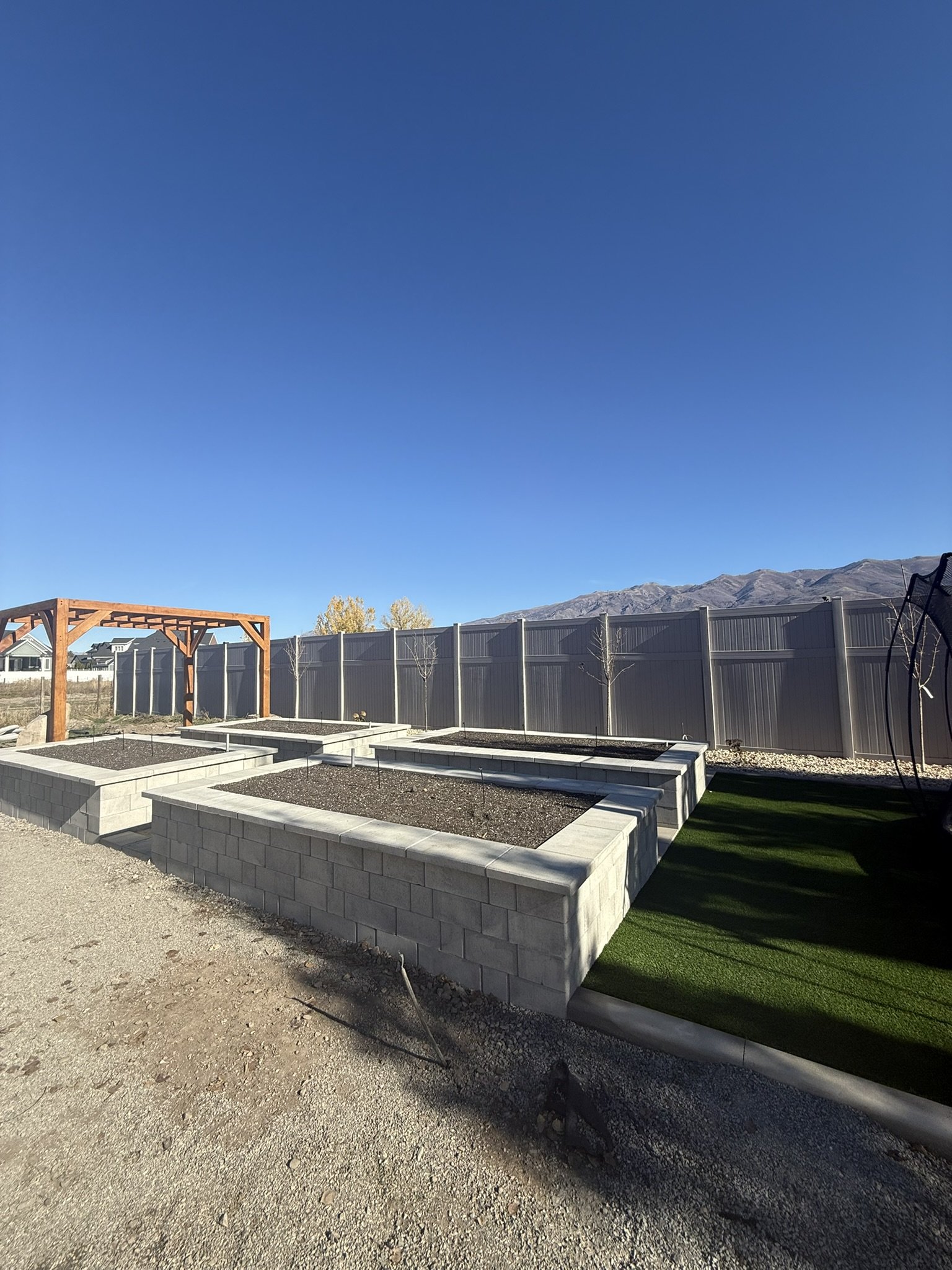 A backyard garden with raised garden beds made of concrete blocks, a wooden trellis on the left side, and a gray fence surrounding the yard. There is a mountain range in the background under a clear blue sky.