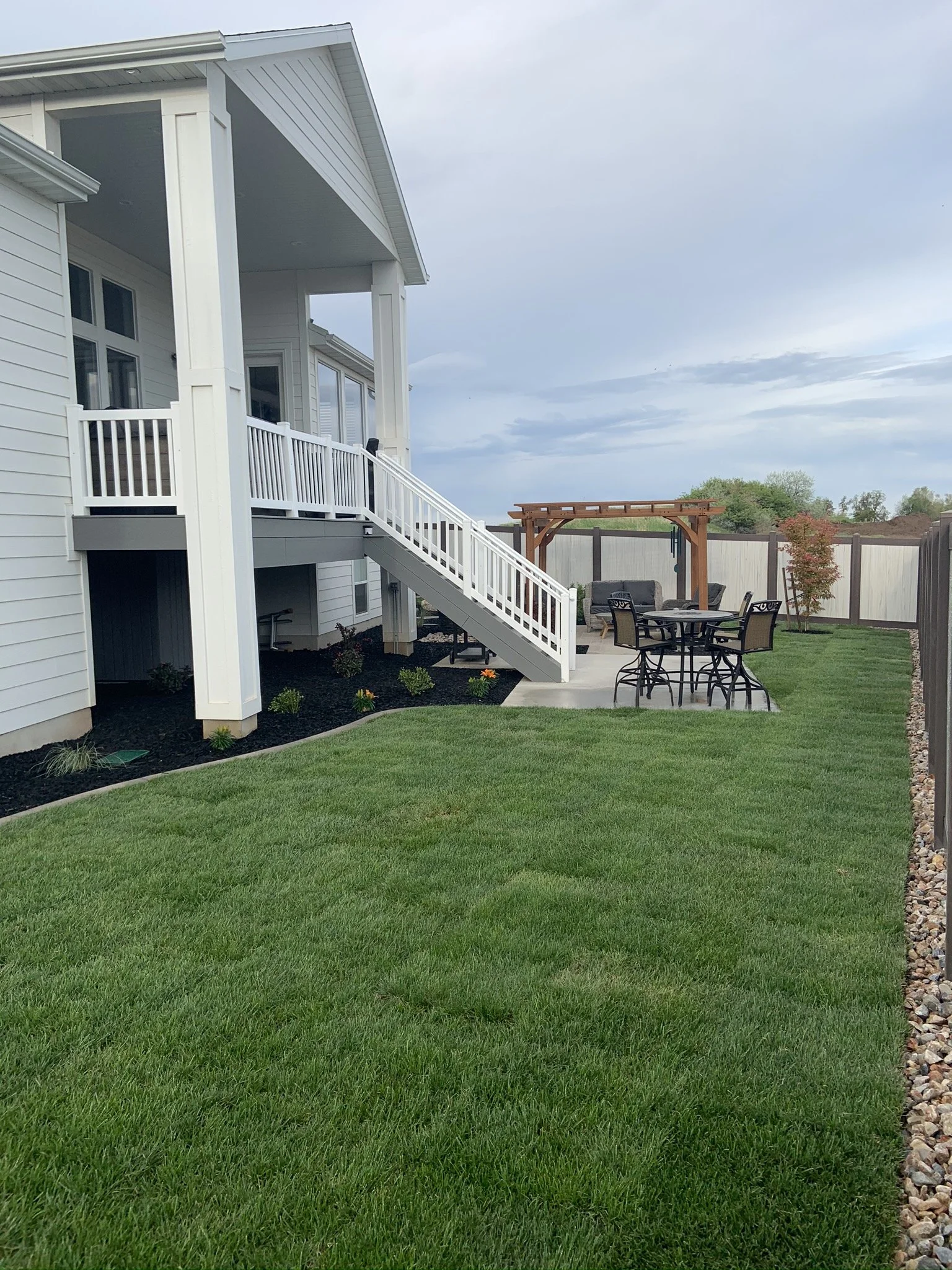 A backyard with a green lawn, a white house with a deck and stairs, and an outdoor patio with seating and a wooden pergola.