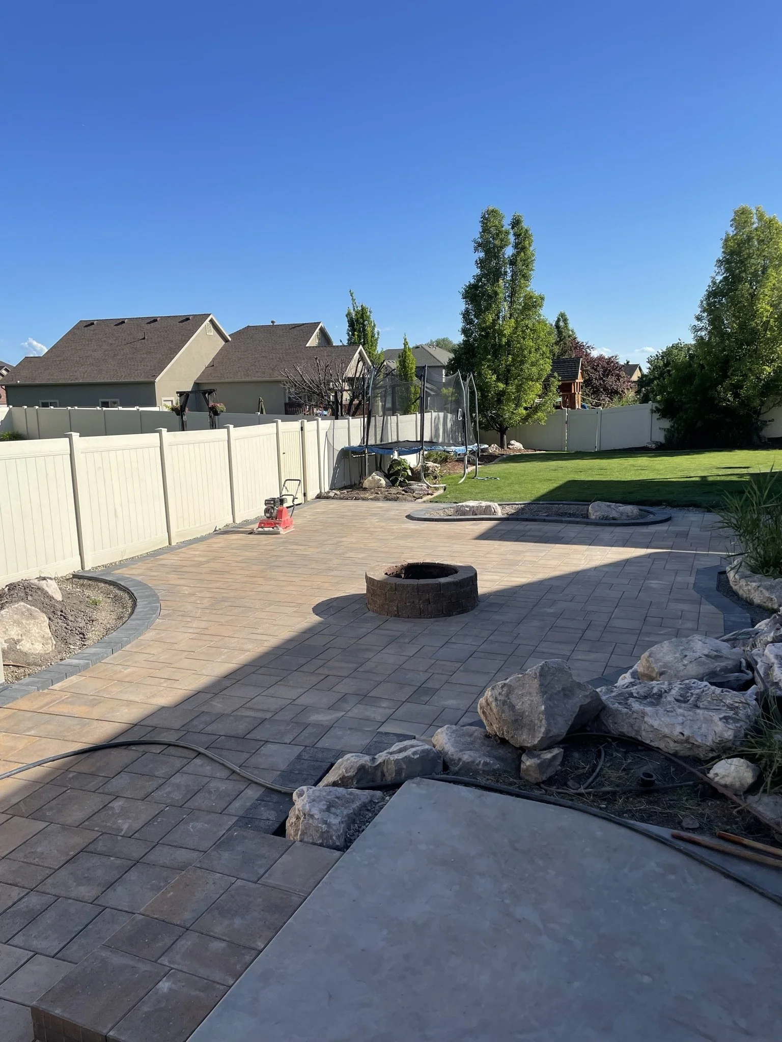 Backyard patio with interlocking pavers, a circular fire pit, landscaped garden beds with rocks and plants, a trampoline, and neighboring houses under a clear blue sky.