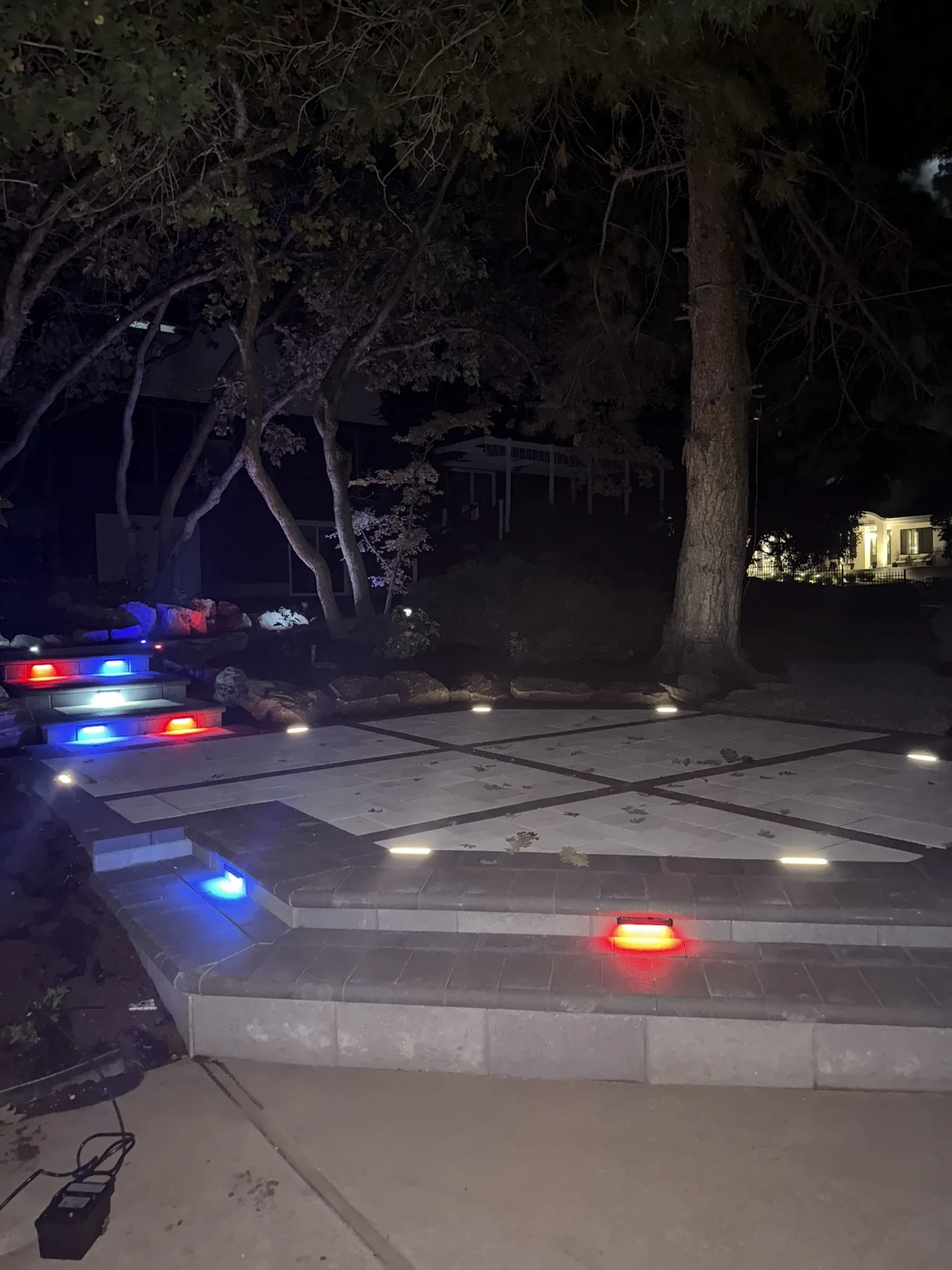 A nighttime view of an outdoor chessboard with illuminated squares and colorful LED lights around its edges, surrounded by trees and a dark yard.