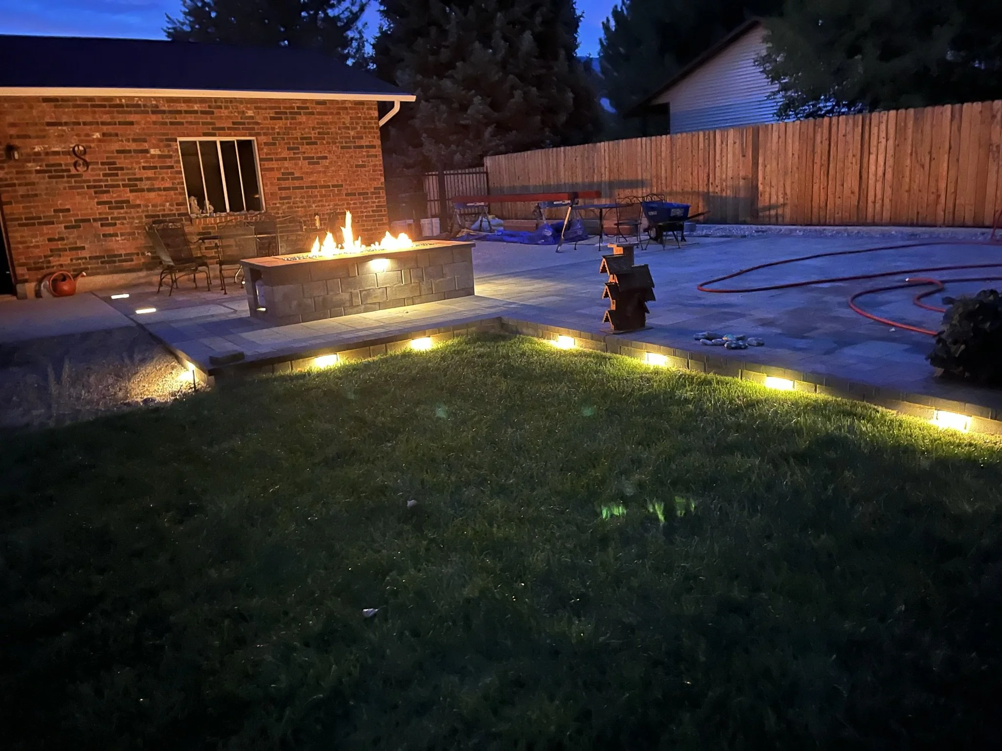 Backyard at dusk featuring a lit fire pit on a patio, illuminated pathway lights along the edge of a lawn, and various outdoor furniture and tools, with a wooden fence in the background.