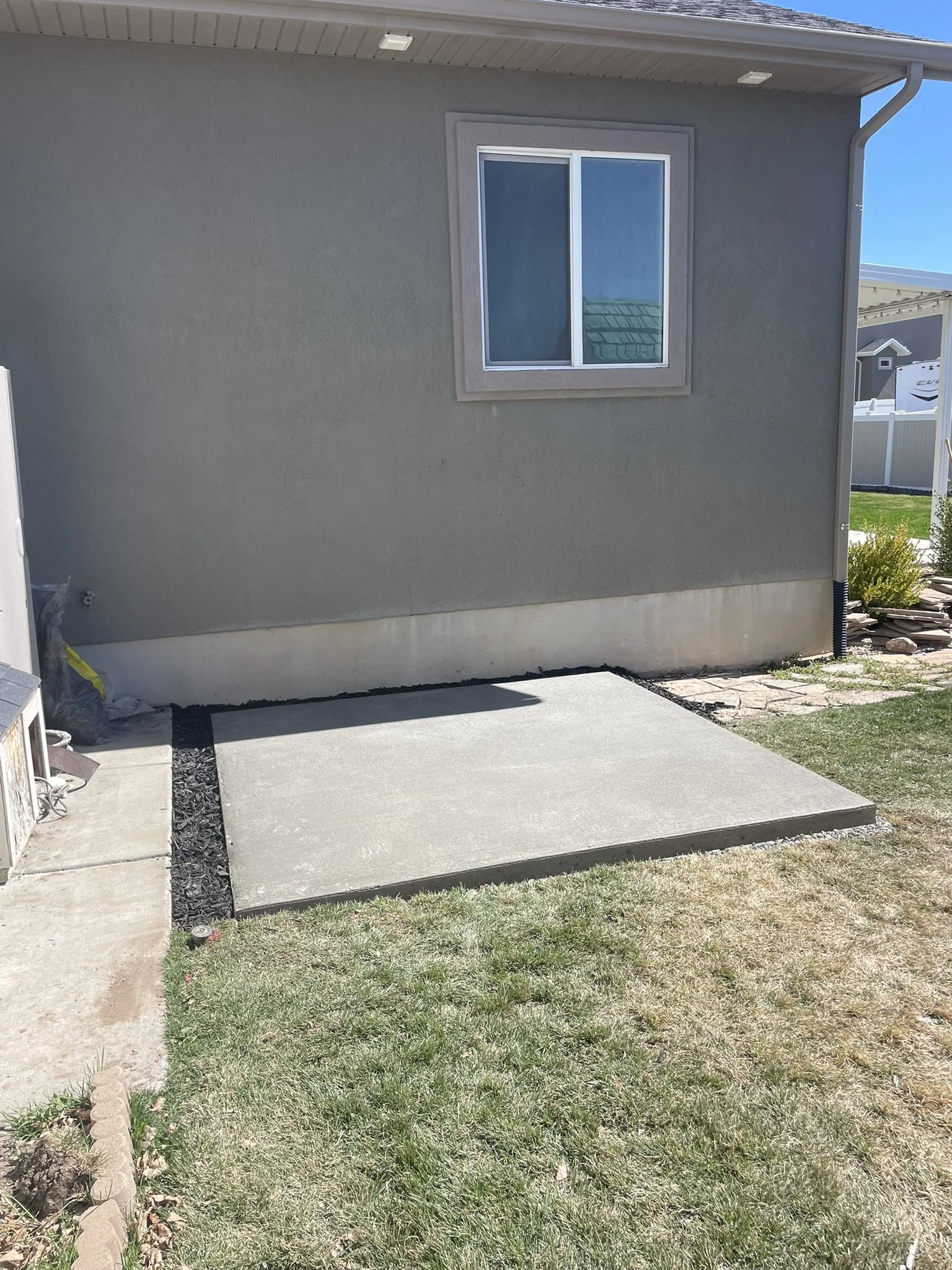 Freshly poured concrete patio next to a house with gray exterior, window, and small front yard with grass.