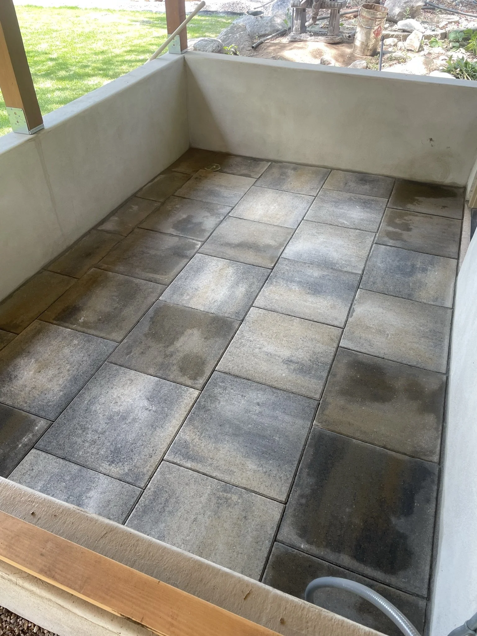A partially enclosed outdoor space with a tiled floor, white wall, and a view of a green backyard with rocks, plants, and gardening tools.