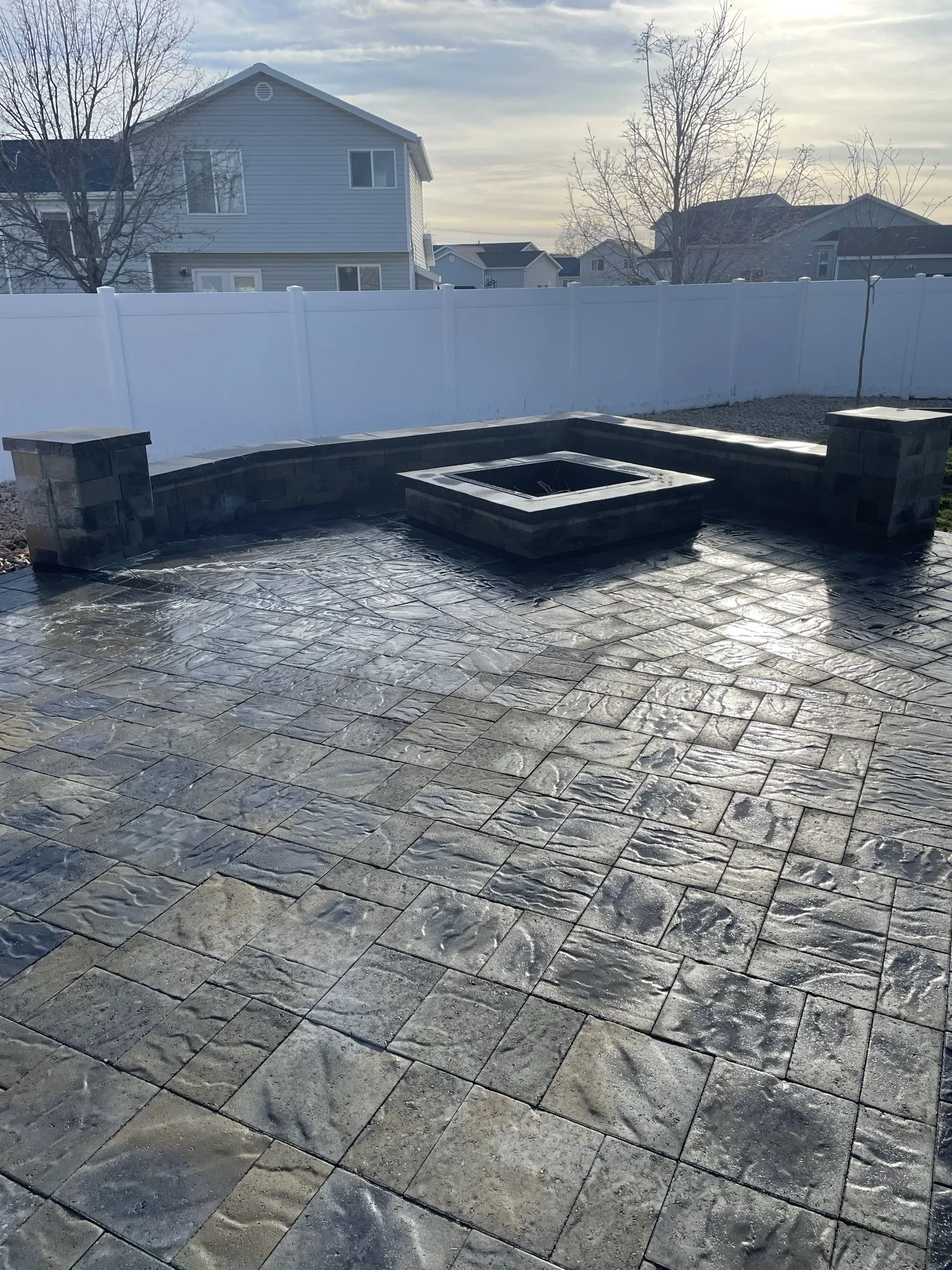 Backyard with concrete patio, small raised fire pit, and two brick columns, enclosed by a white fence, with neighboring houses and trees in the background.