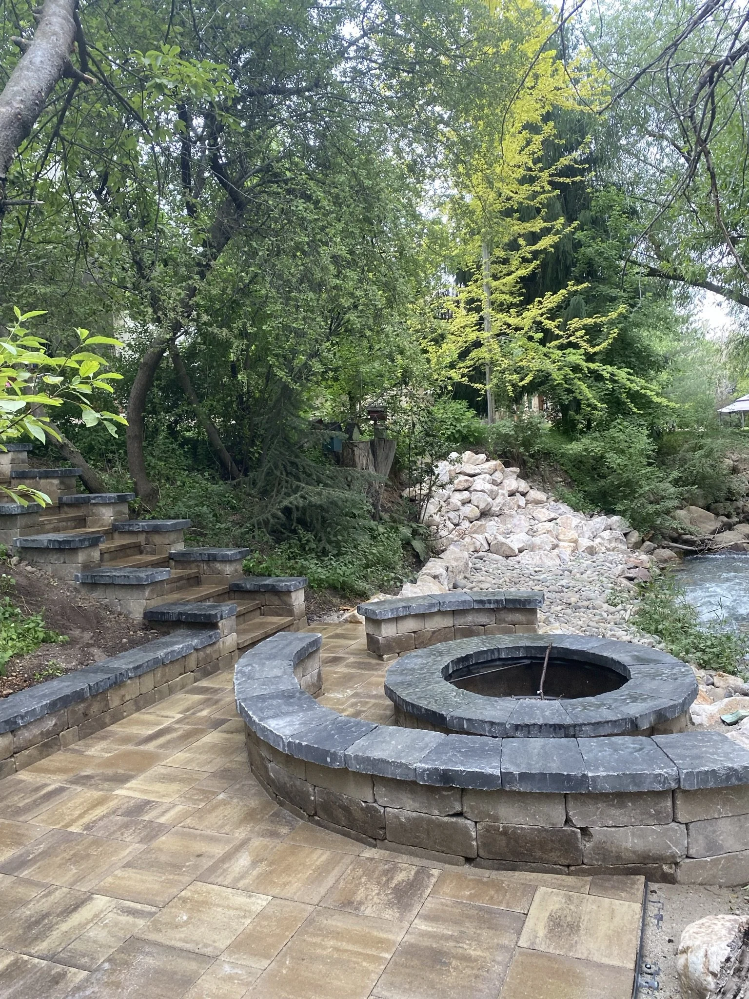 A patio next to a river with stone steps, a circular fire pit, and lush green trees.