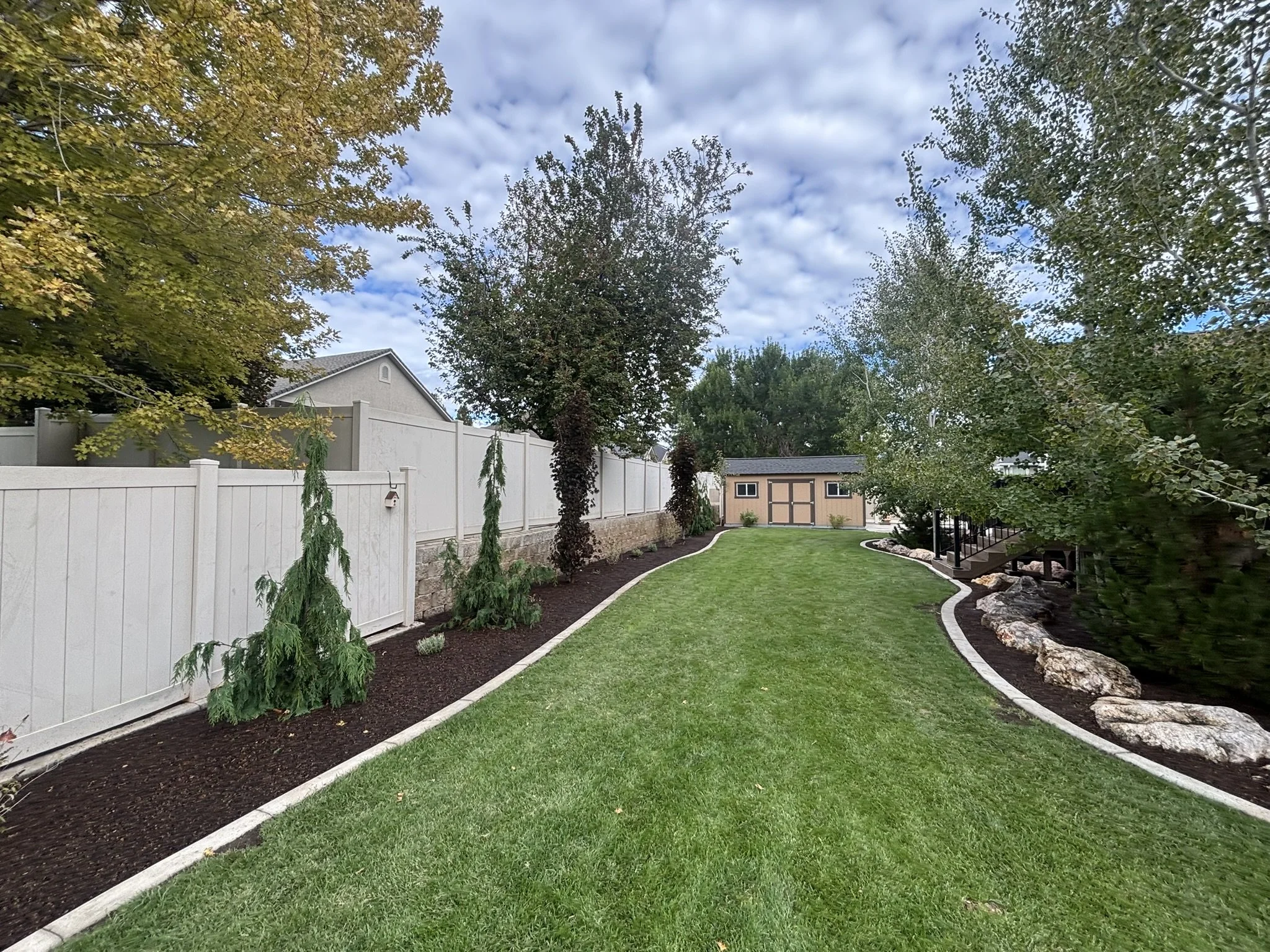 A backyard with a lush green lawn, curved flower beds with mulch, white decorative stone borders, and trees along the fence. There's a small shed in the background and a staircase on the right side.