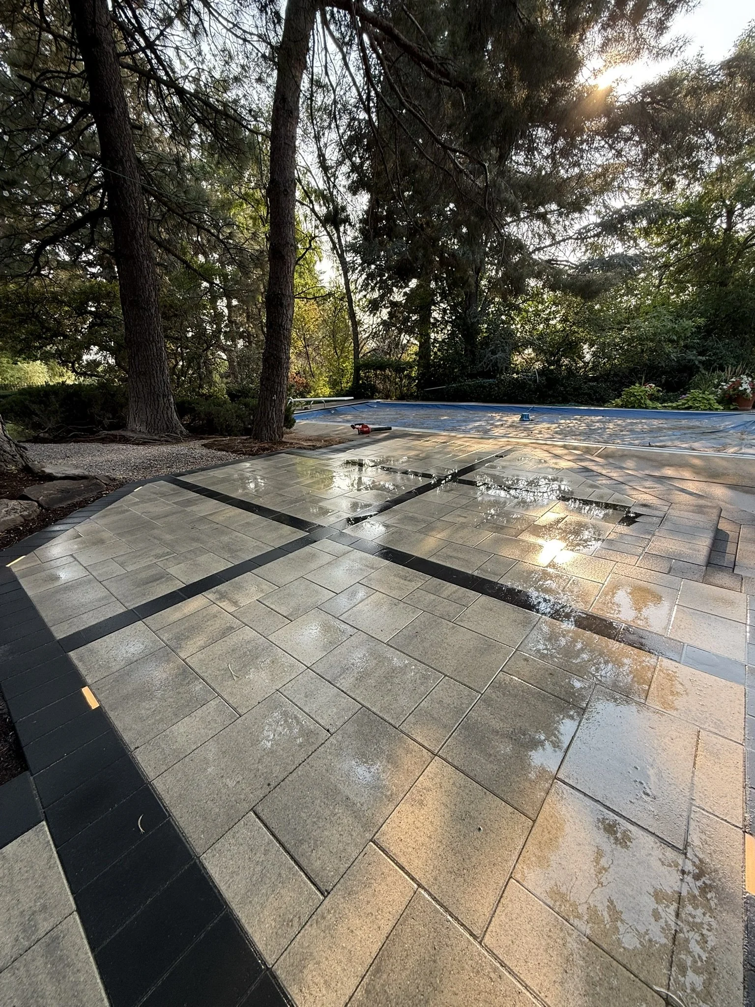 Wet outdoor tiled patio with trees in the background, sunlight filtering through the leaves, and gardening tools on the ground.
