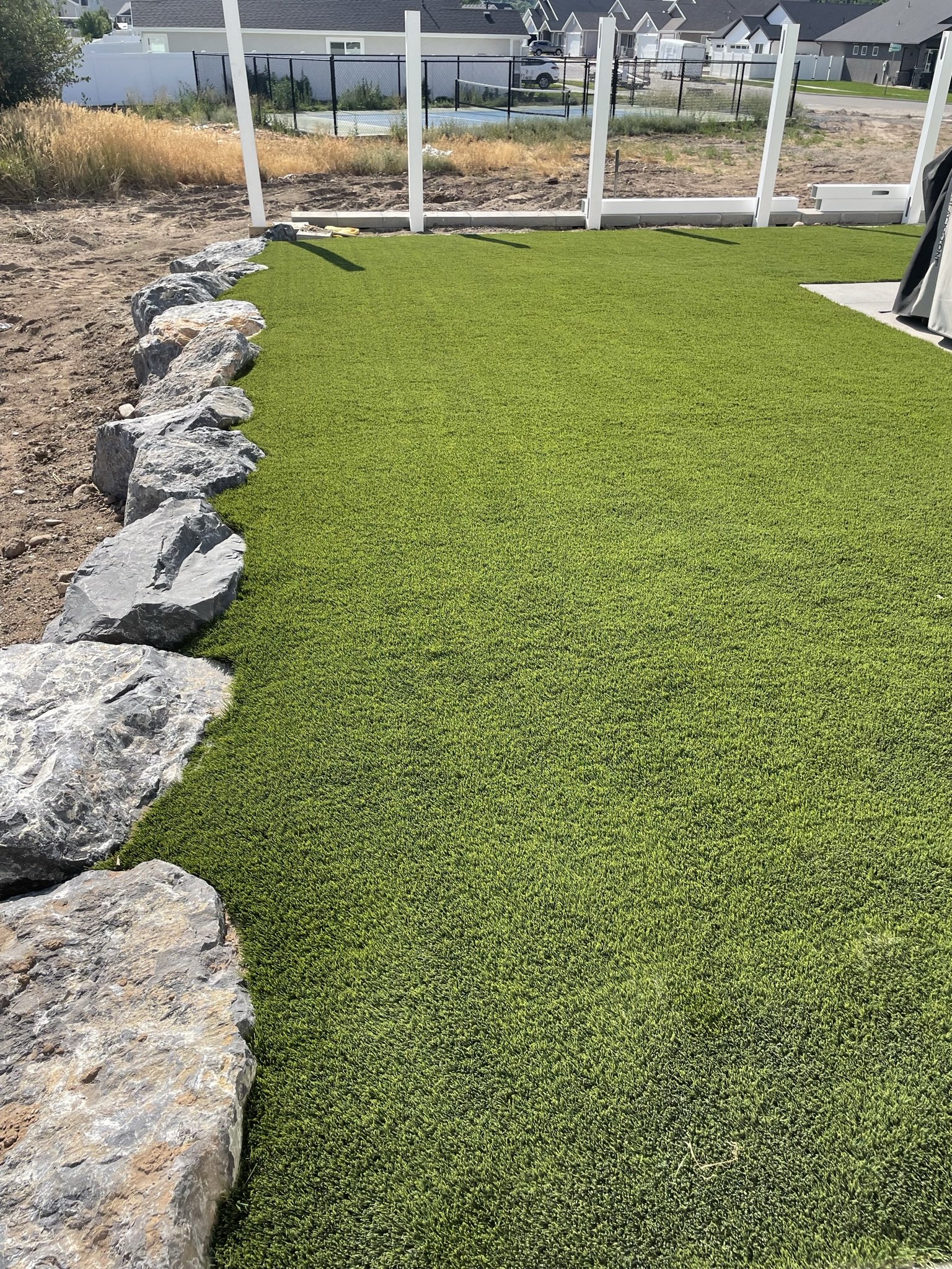 A backyard with a freshly laid green artificial turf lawn bordered by large rocks on the left side and a white fence in the background.