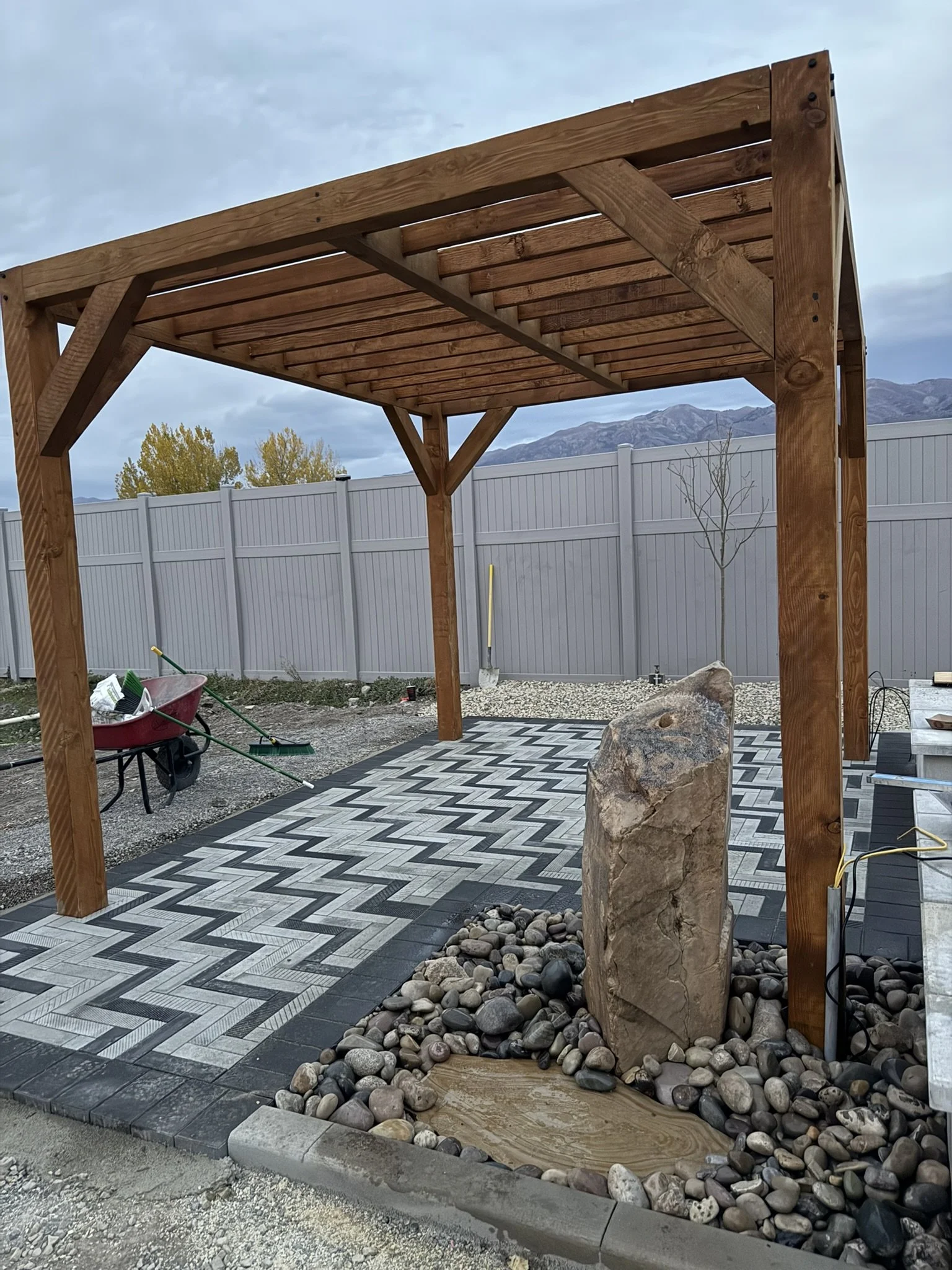 A backyard patio with a wooden pergola, patterned paving stones, a large rock fountain, and decorative pebbles, with a gray fence and mountains in the background.