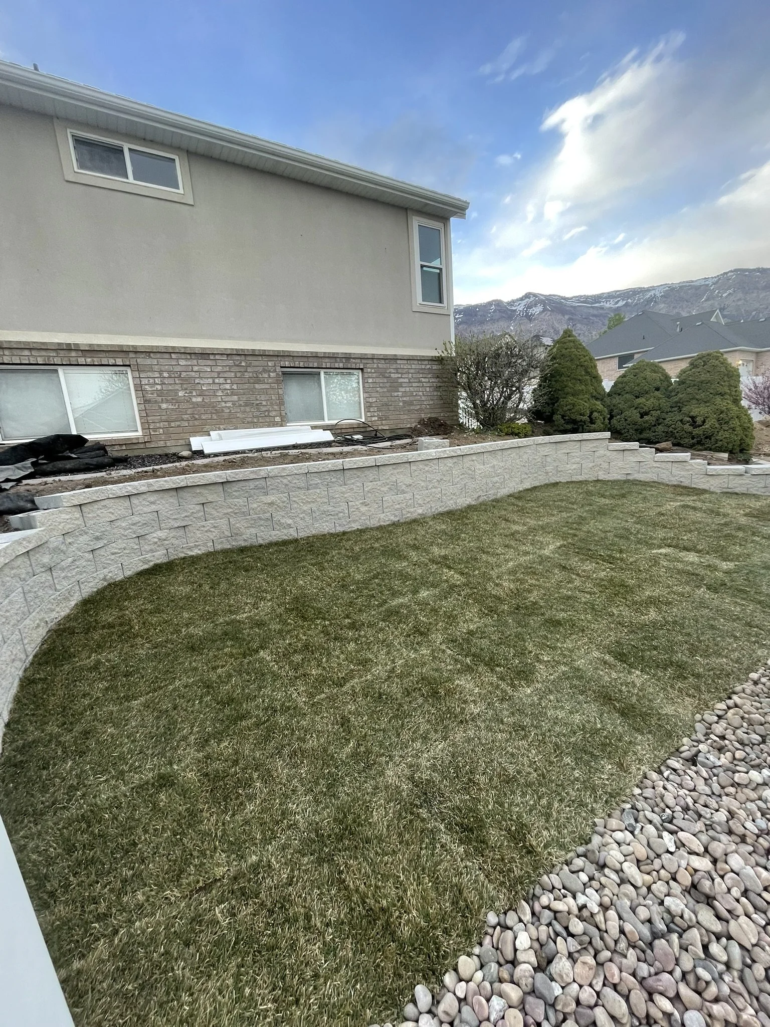 A backyard with a curved stone retaining wall, freshly planted grass, and a row of small rocks along the edge. In the background, there's a two-story house with a brick and beige siding exterior, windows, and a mountain range under a partly cloudy sk