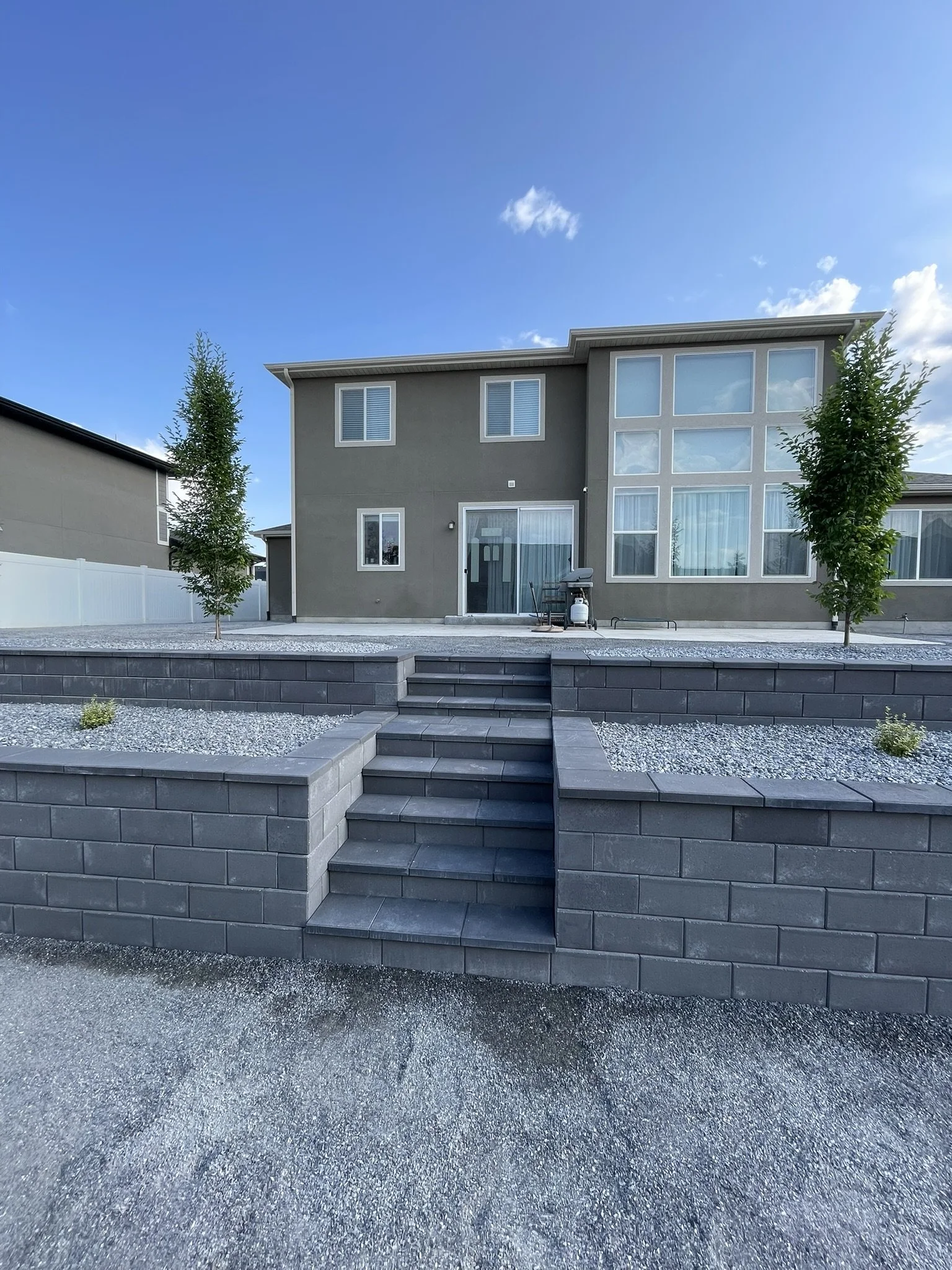 Back view of modern gray two-story house with large windows, small trees, and a tiered yard with steps, gravel, and a patio with a grill.