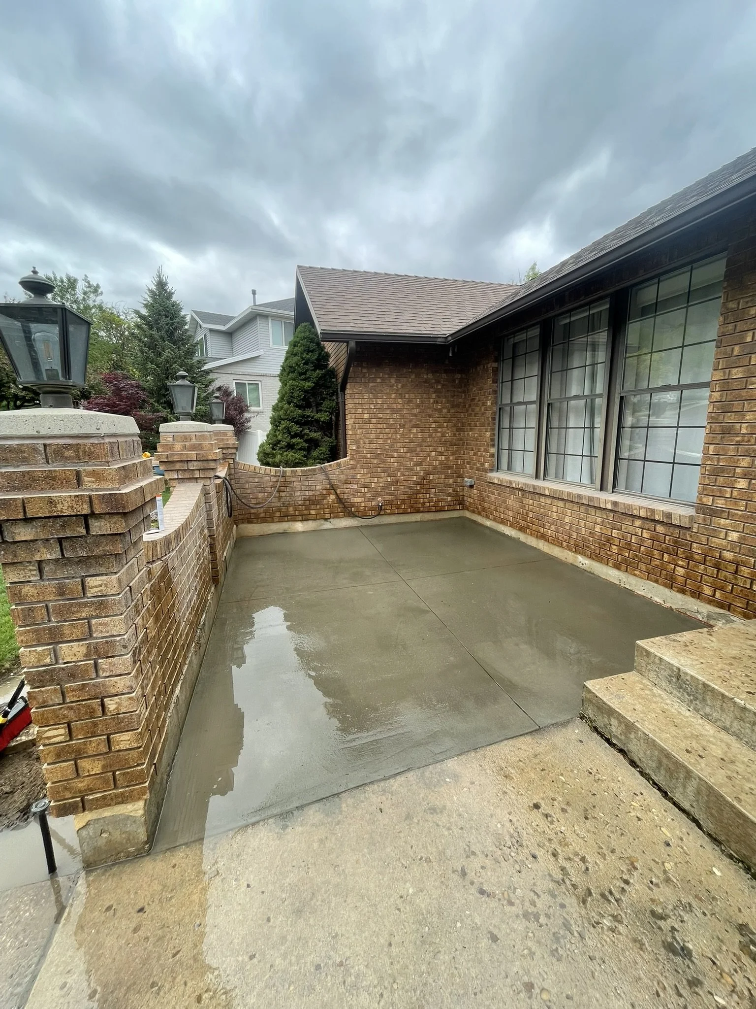 Wet concrete patio outside a brick house with large windows, a brick wall, and outdoor lamps, under a cloudy sky.