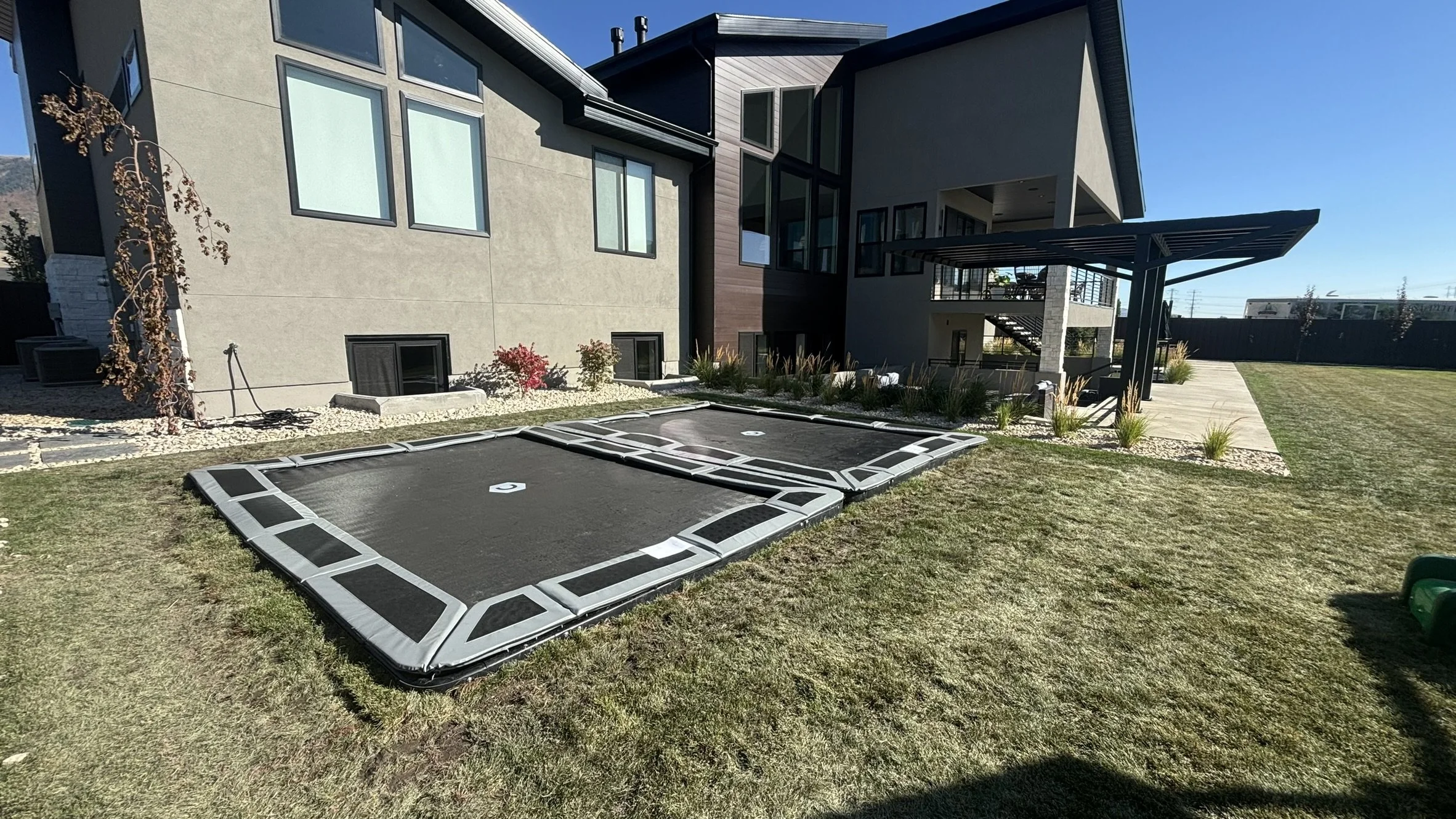 Modern house with two mini trampolines set in the backyard, green grass, and a patio with outdoor steps leading to a second-story balcony.