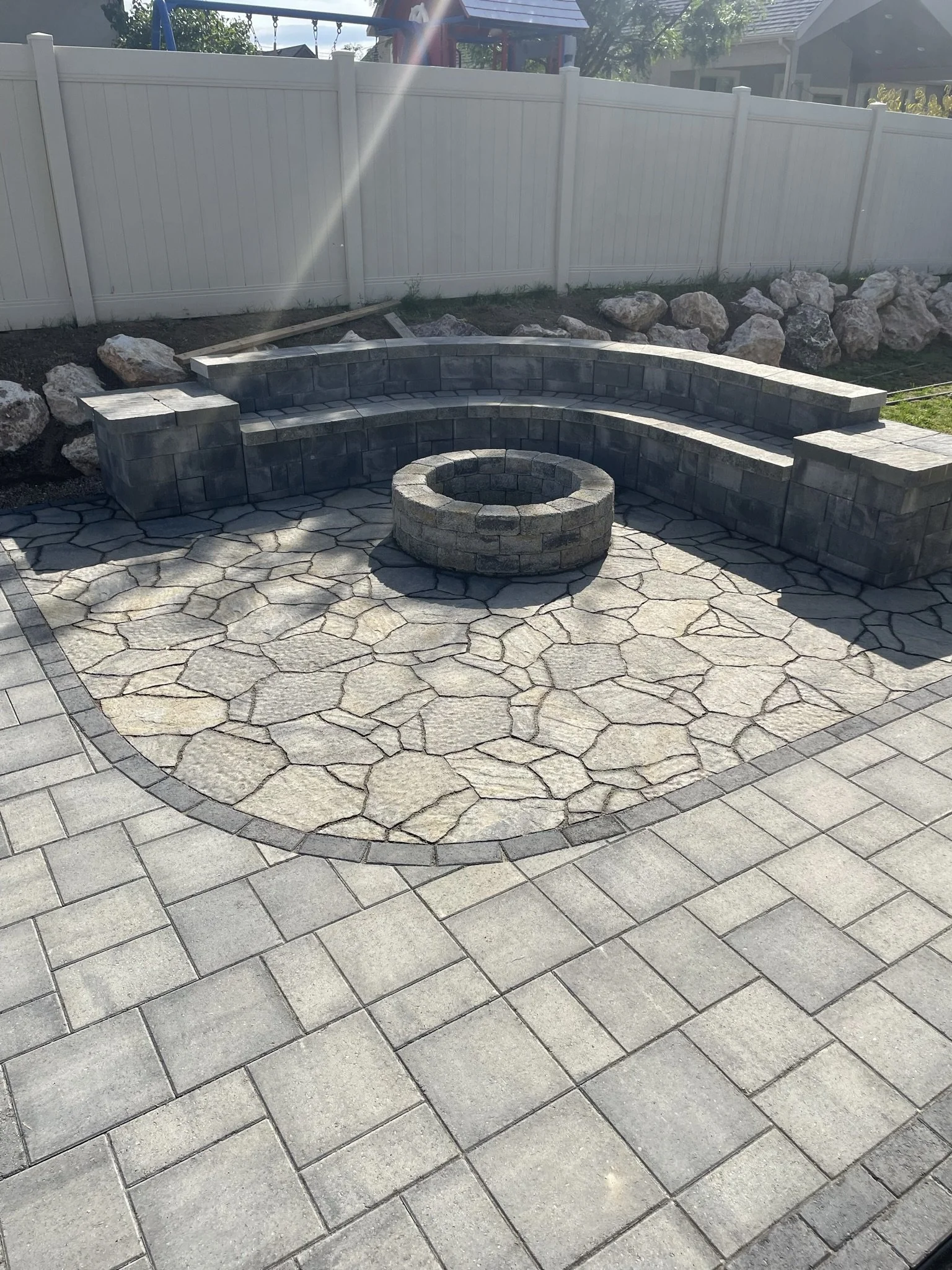 Backyard featuring a stone fire pit surrounded by a curved stone seating area and stone patio with paver borders, enclosed by a white fence with rocks and a playset in the background.