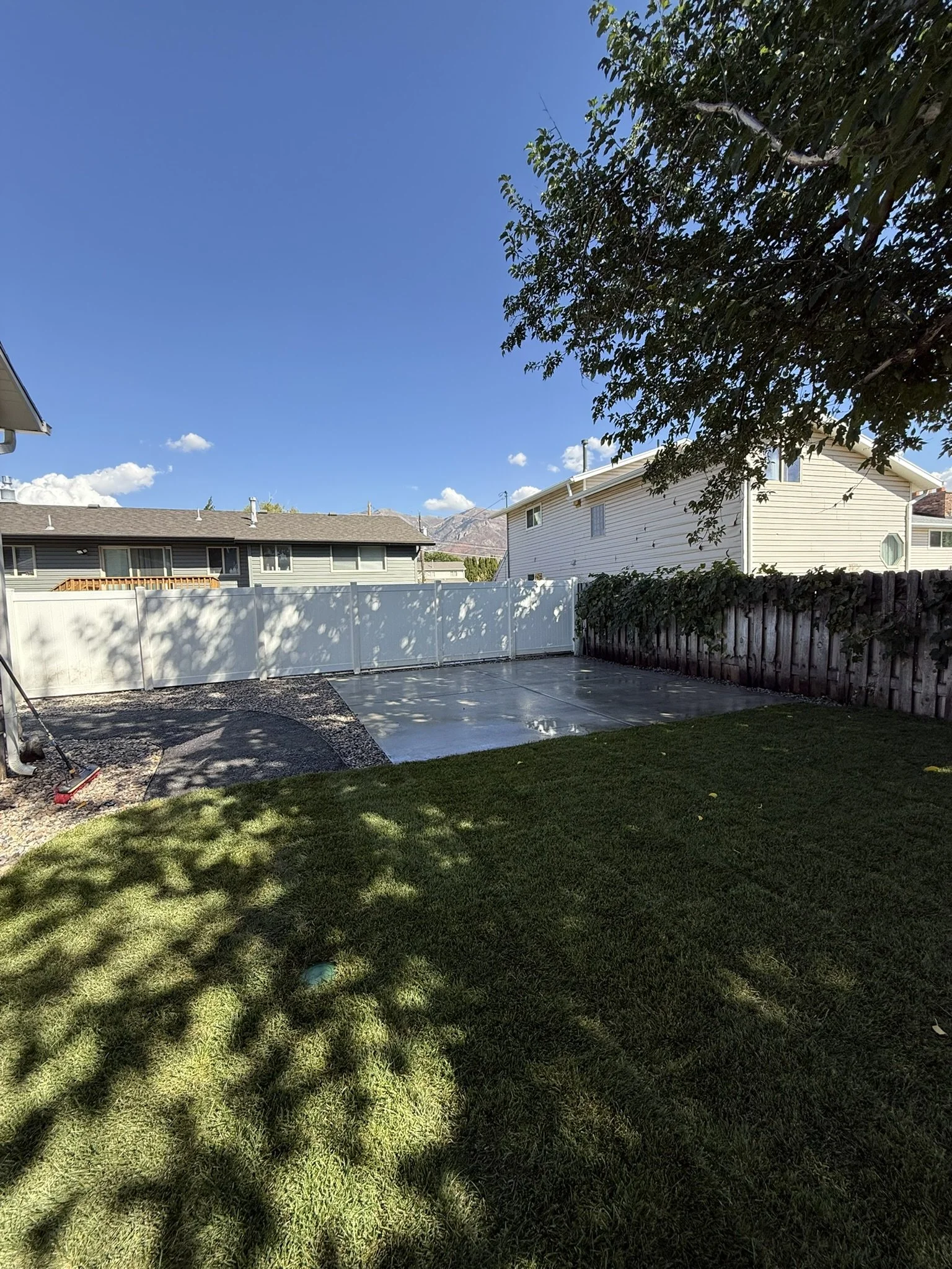 Backyard with a patch of green grass, a concrete patio, a white fence, and a wooden fence, with a tree casting shadow on the grass and a clear blue sky with a few clouds.