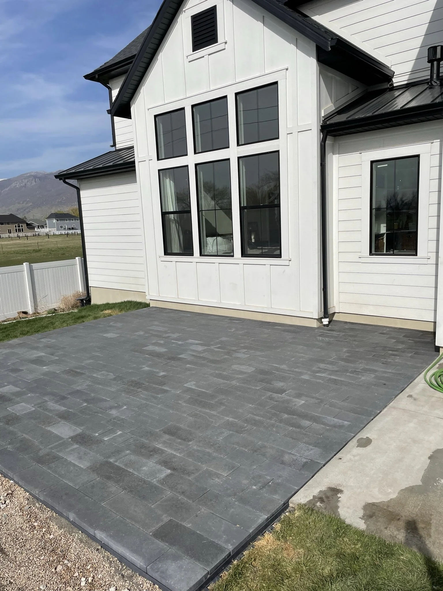 A newly paved brick patio outside a modern white house with large black-framed windows and a dark roof, with a view of a rural landscape and mountains in the background.