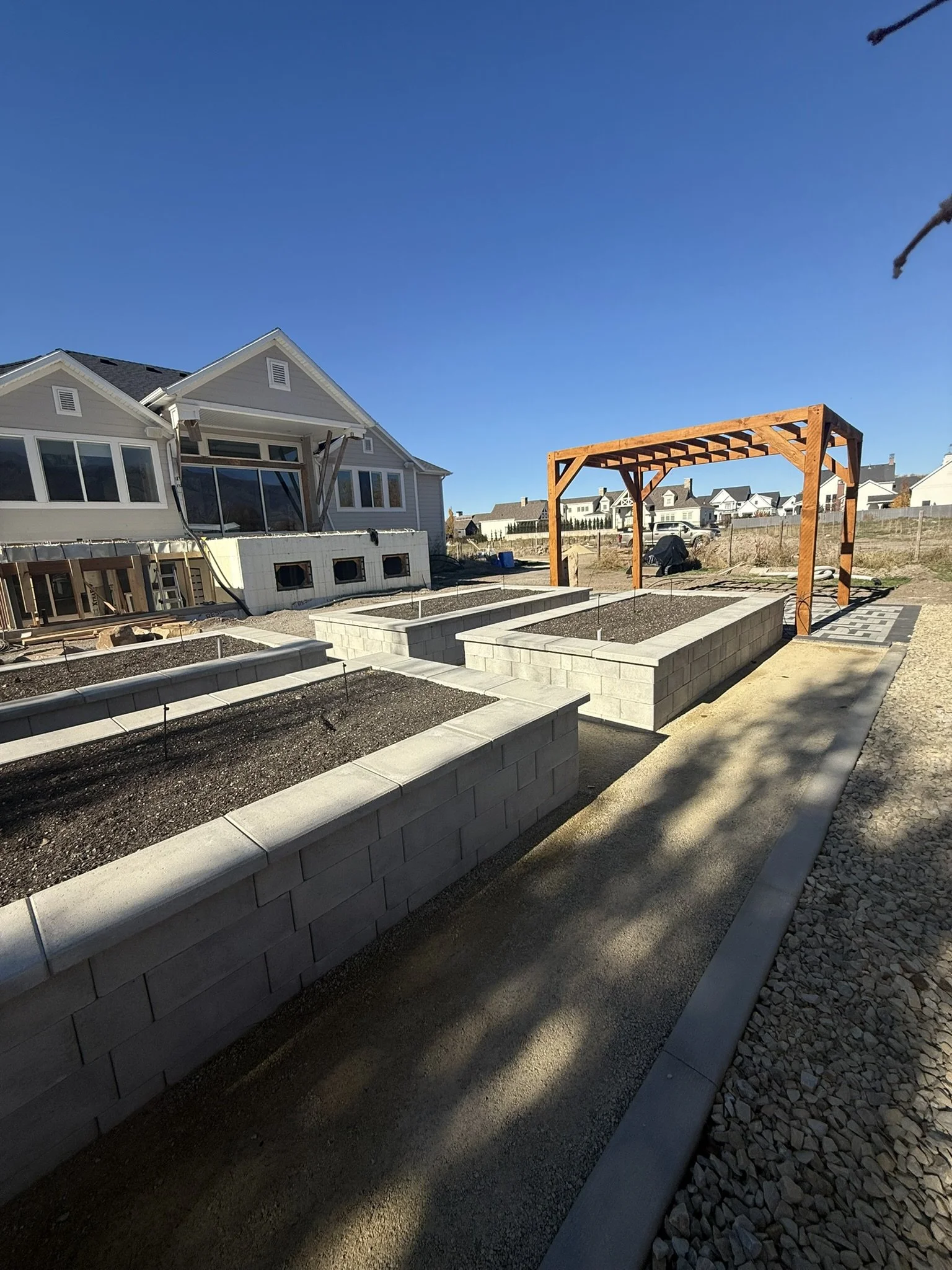 A backyard with raised garden beds constructed from concrete blocks under a wooden pergola, with a house in the background under a clear blue sky.