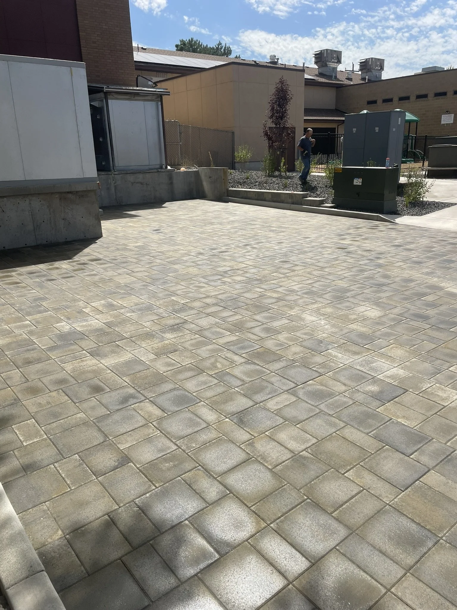 An outdoor area with a paved surface made of beige and gray bricks, near a building with utility boxes and a small landscaped section with plants and a small tree. There is a man walking in the background.