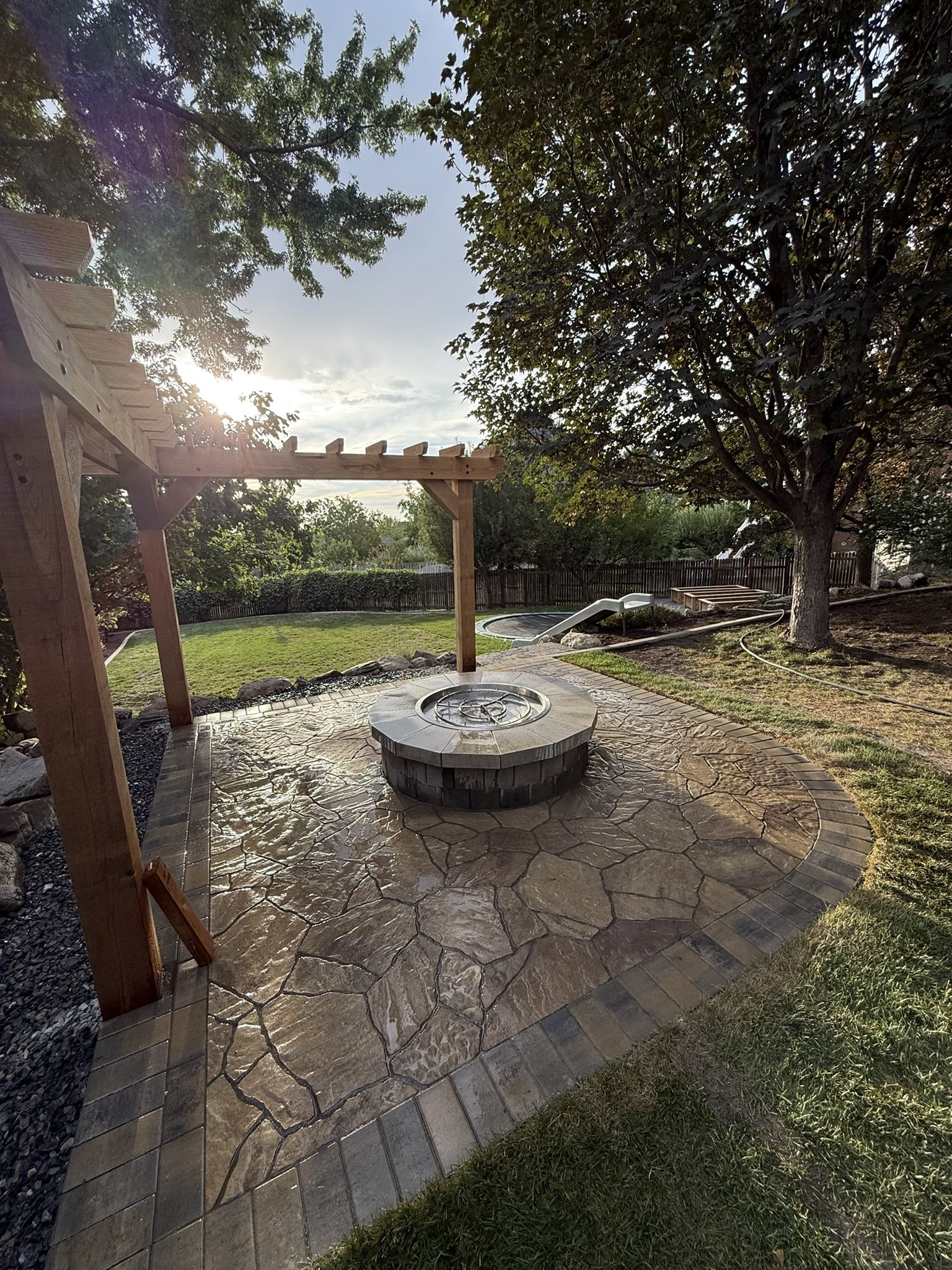 A backyard with a stone paved patio, a wooden pergola, a fire pit, a grassy area, trees, and a small slide in the distance.