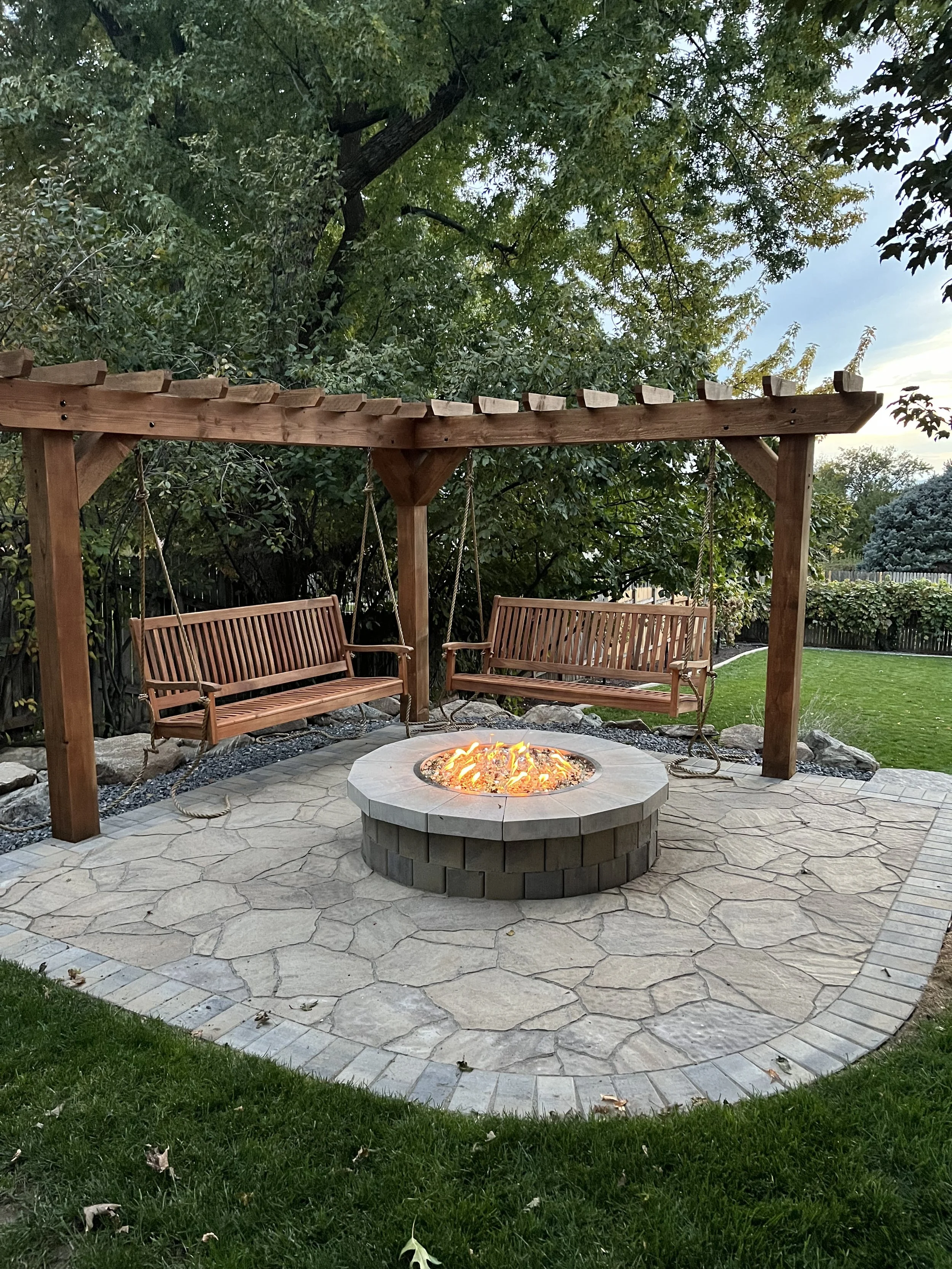 Backyard patio with a fire pit in the center, two wooden swings hanging from a wooden pergola, stone pavers, green grass, and trees in the background.