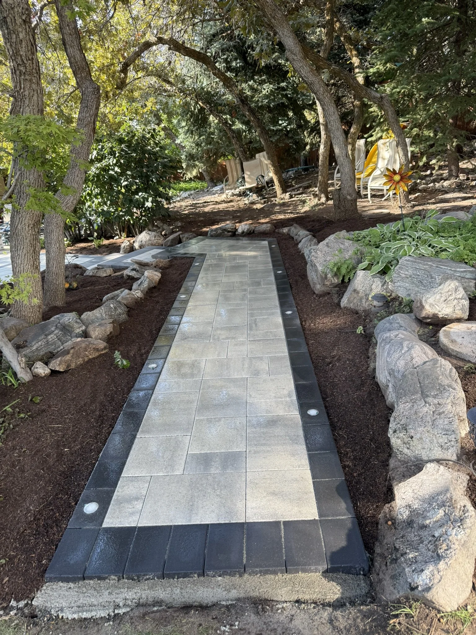 A landscaped backyard with a narrow paved walkway bordered by rocks and mulch, surrounded by trees and shrubs, with a garden flower and outdoor chairs in the background.