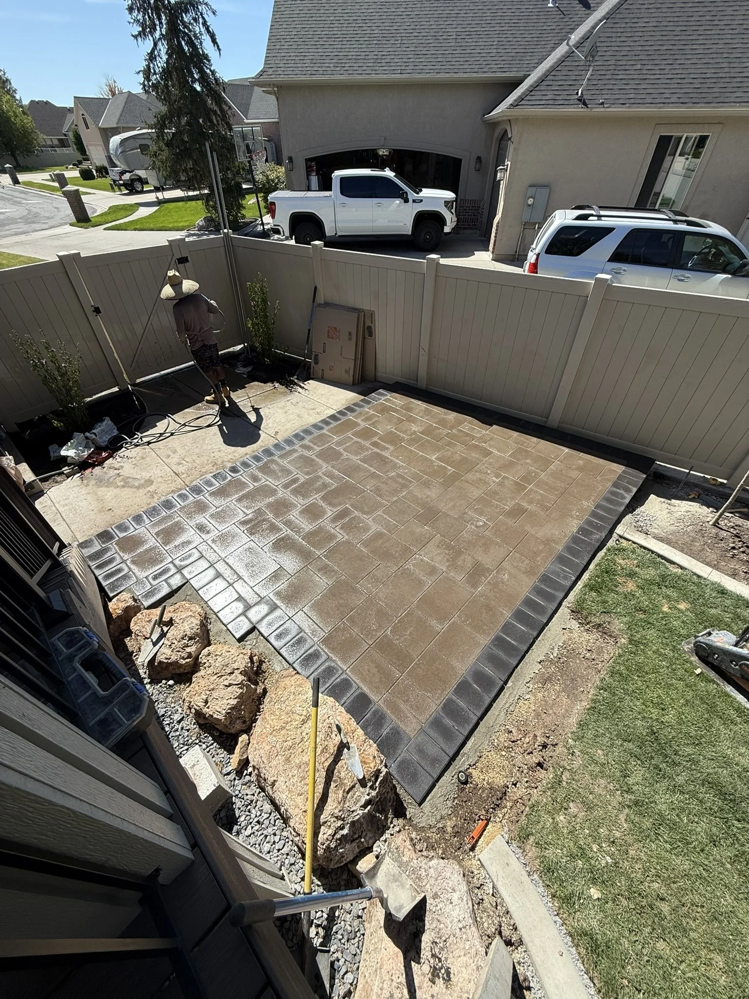 Person installing or sealing pavers in a backyard patio, with several large rocks and construction tools nearby, and a white fence surrounding the yard.