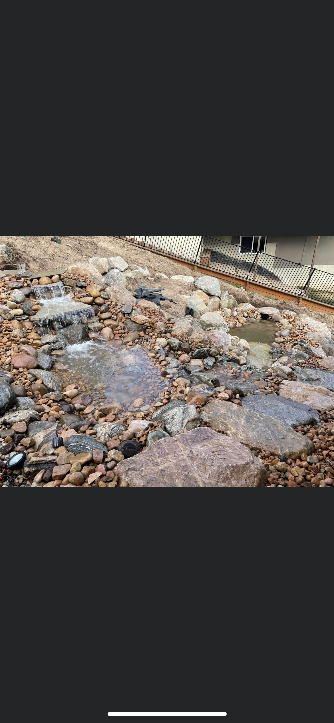 A landscaped backyard with a small multi-tiered waterfall and a pond surrounded by rocks, with a house and a fence in the background.