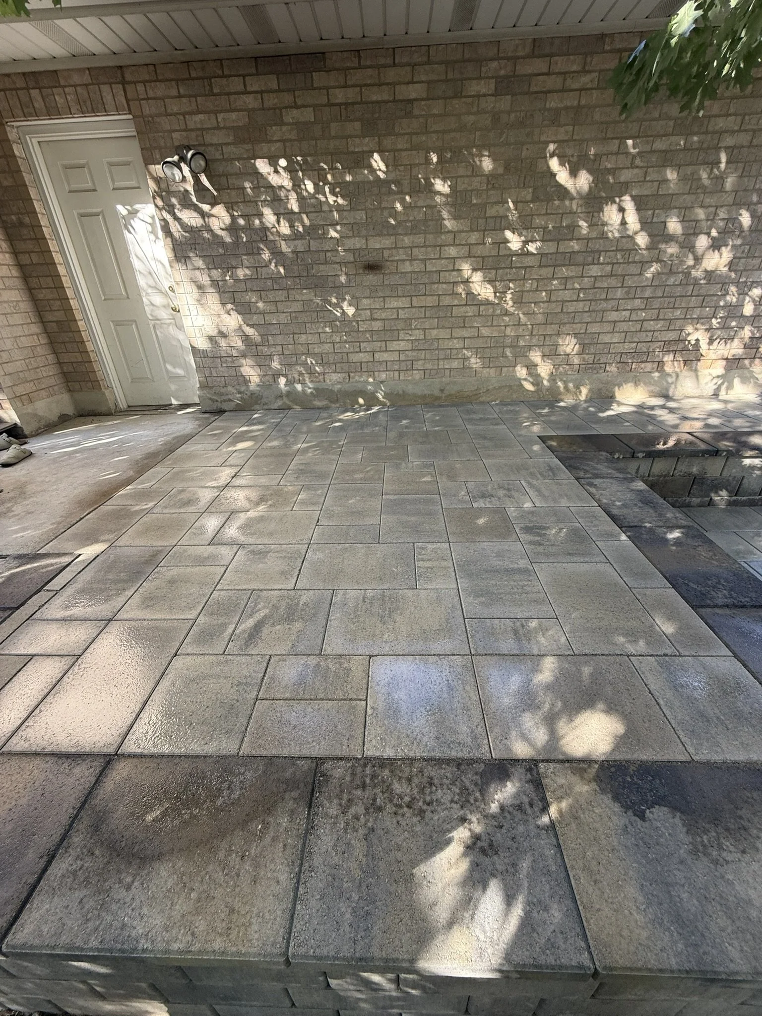 Patio with new rectangular stone pavers, partial concrete area, brick wall with door, and some shadow patterns from tree branches.