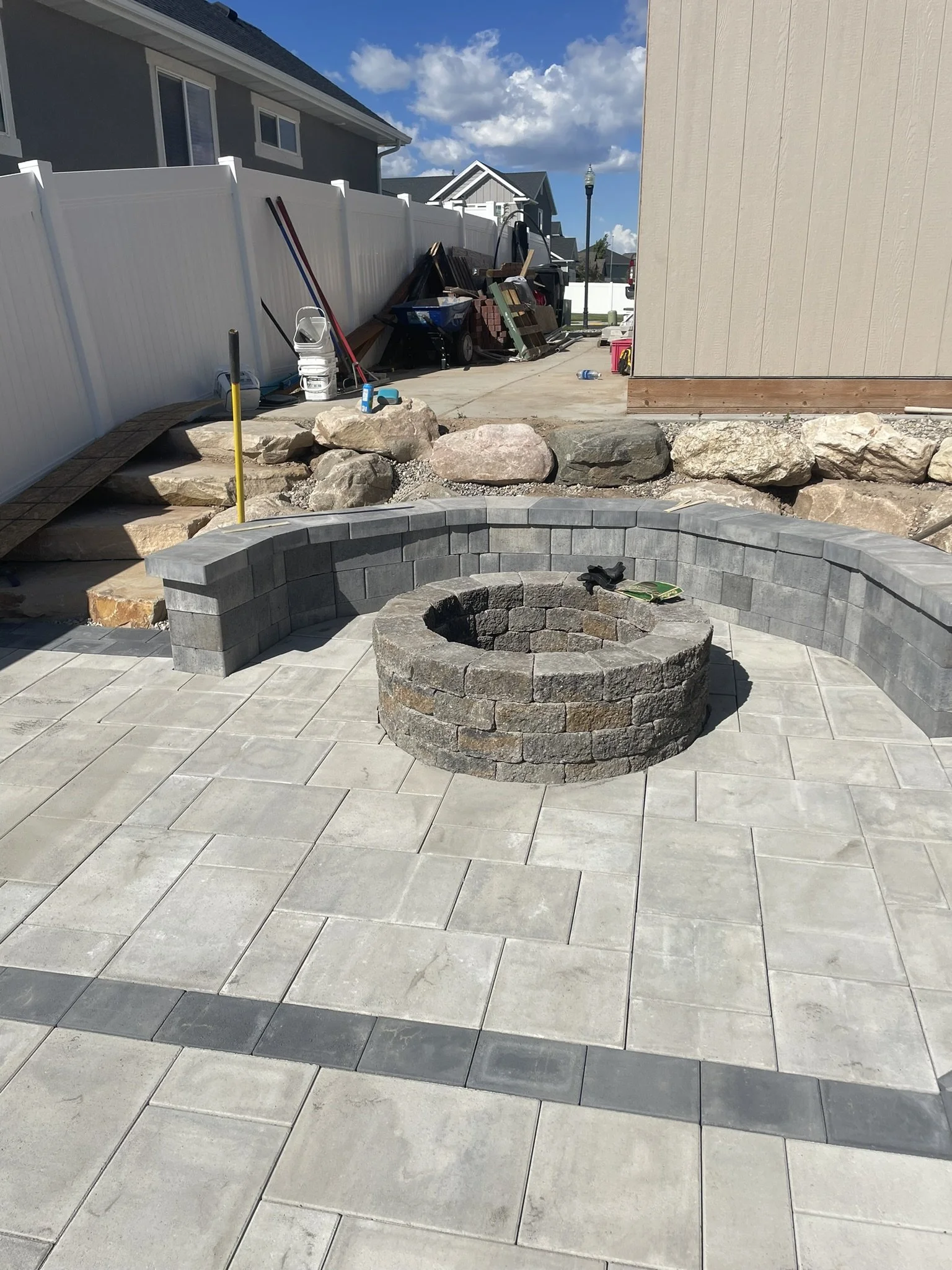 Backyard patio with a circular fire pit made of dark gray stone, surrounded by gray concrete pavers. A curved stone bench built into a low wall is seen behind the fire pit. In the background, construction materials and tools are scattered, including 