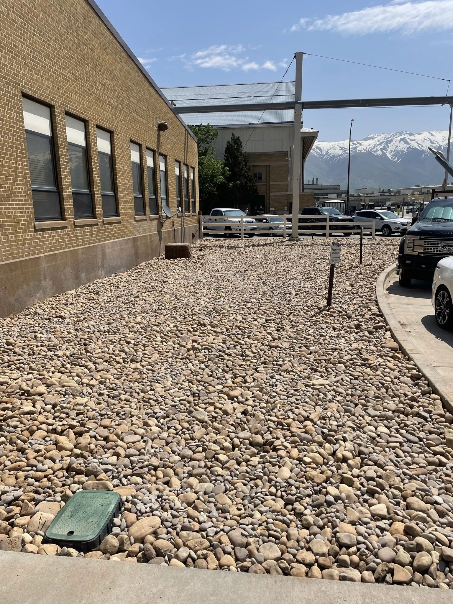 A parking area with a bed of small, rounded stones, a parking sign, and a row of cars, with mountains in the background and a partly cloudy blue sky.