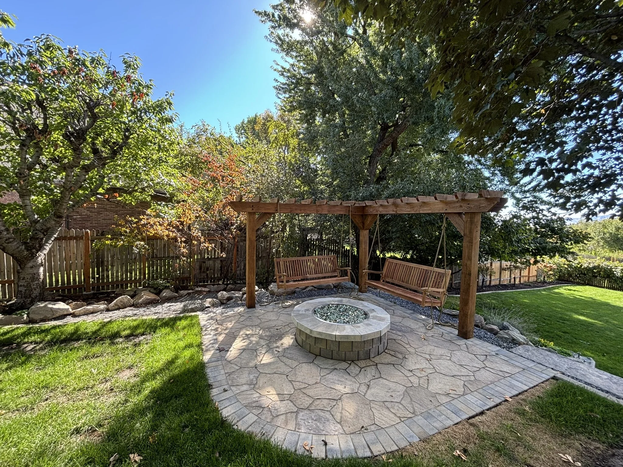 A backyard patio with a fire pit, two wooden swing benches hanging from a wooden pergola, surrounded by green grass, trees, and a wooden fence under a blue sky.