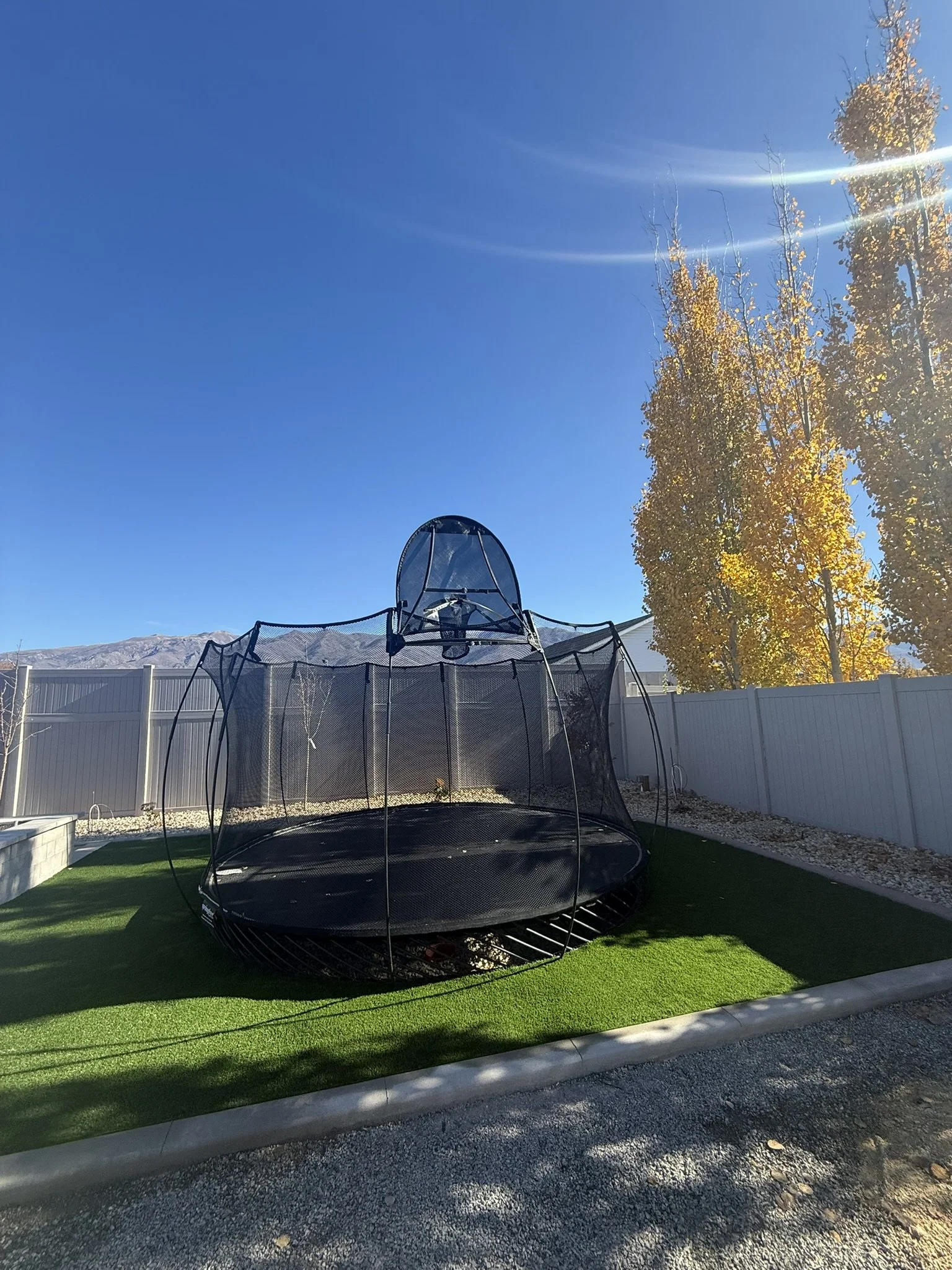 Backyard with a trampoline, artificial grass, white fence, trees with fall leaves, and mountain in the distance under a clear blue sky with sunlight.