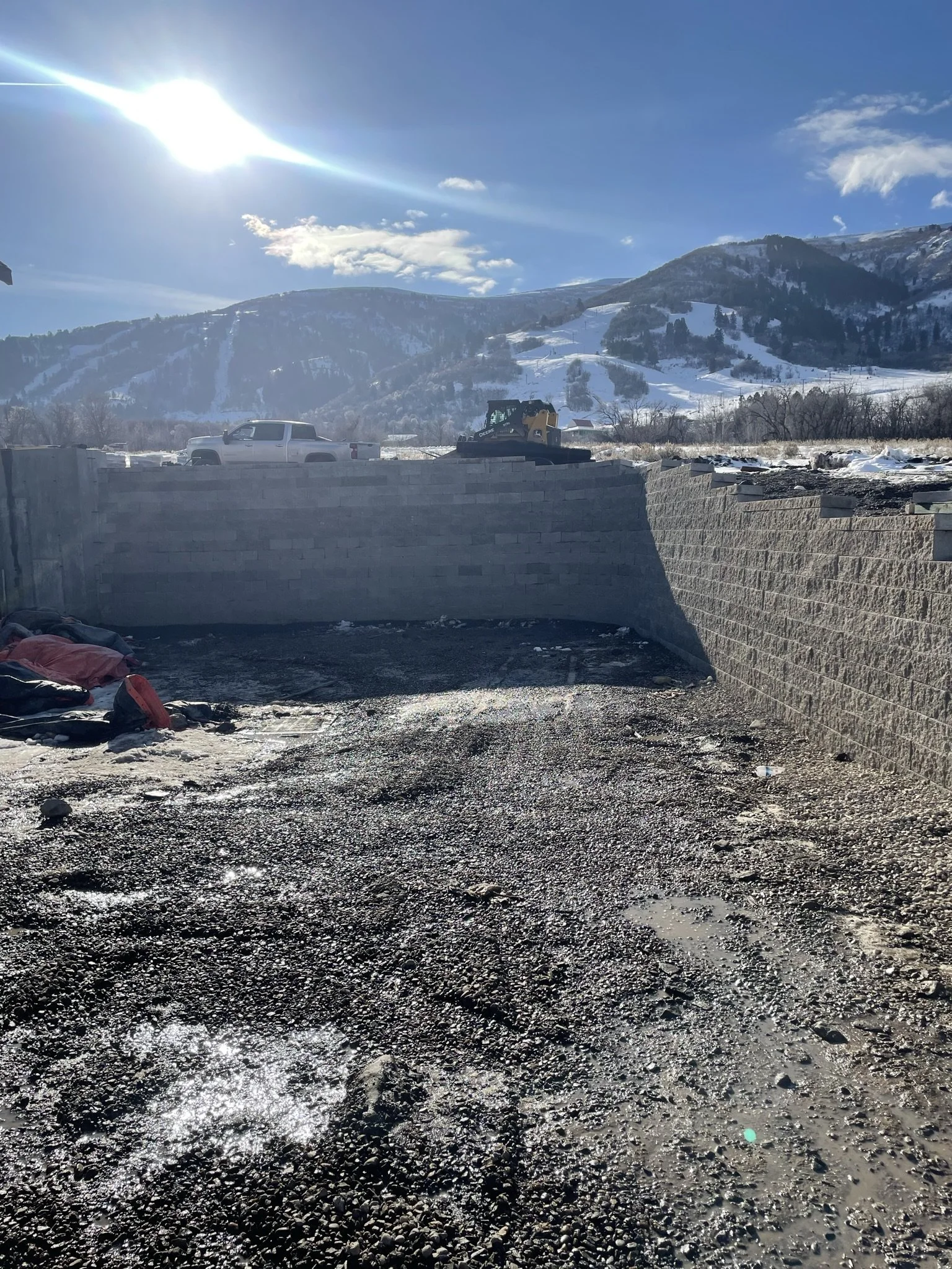 Empty outdoor construction site with gravel ground, partially built concrete block wall, snow in the background, and mountains under a bright sunny sky.