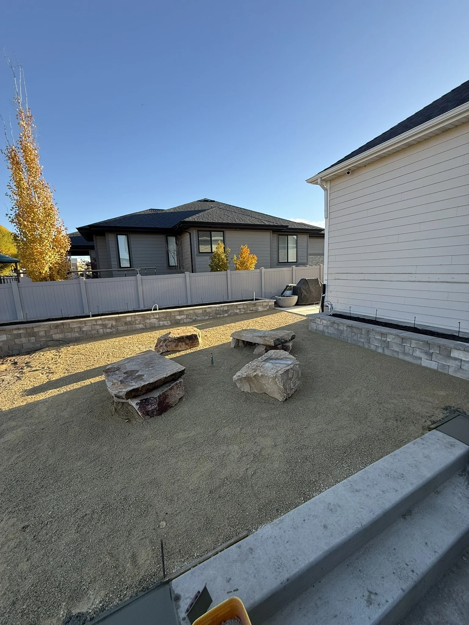 Backyard with sand and four large rocks, partially constructed retaining walls, neighboring houses, and a clear blue sky.