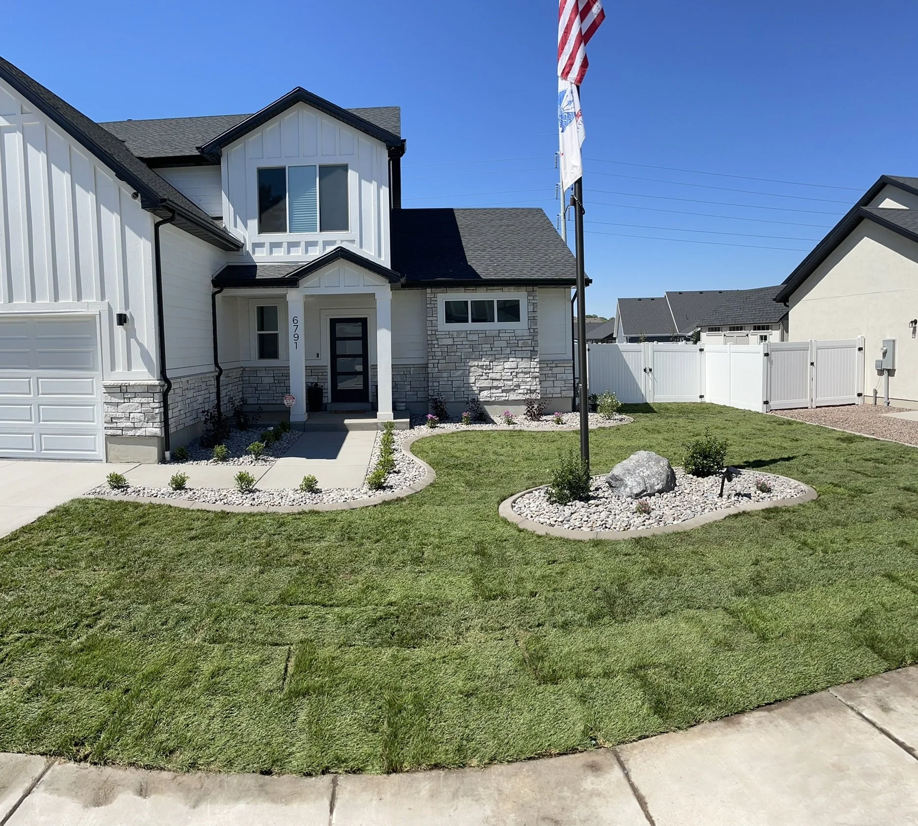 Front yard of a modern house with green grass, a small flower bed with rocks, and a flagpole with American and other flags. White fencing surrounds the yard, and the house features white siding and stone accents.