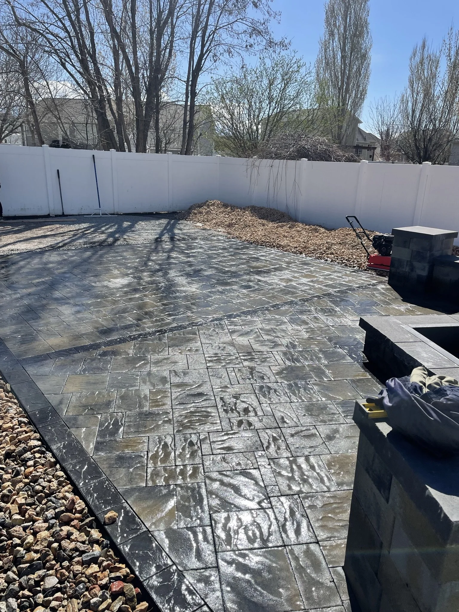 A backyard with newly installed stamped concrete patio, surrounded by a white fence and trees, some with budding leaves.
