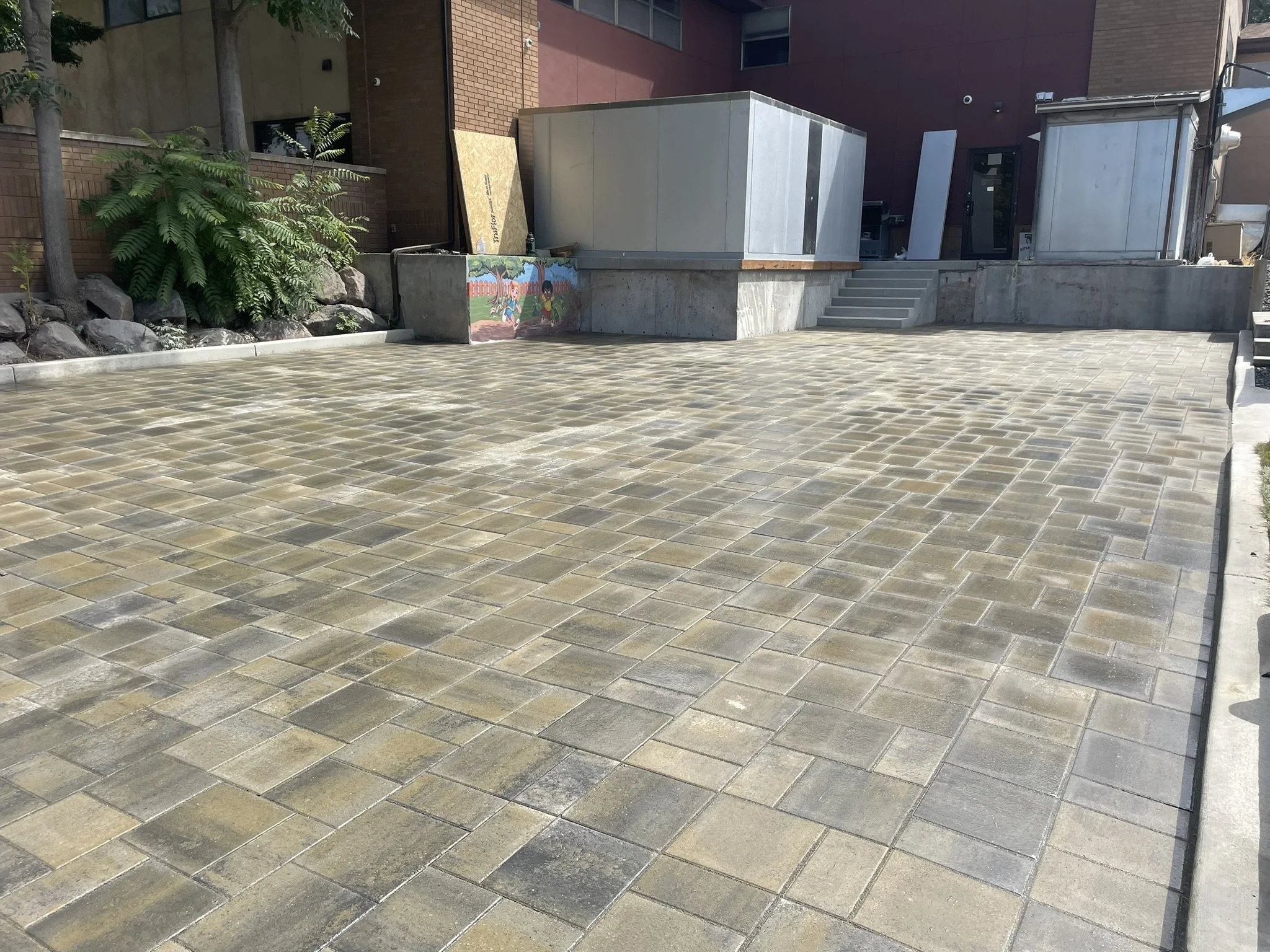 Empty paved outdoor courtyard with square stone tiles, green plants on the left, and building structures in the background.