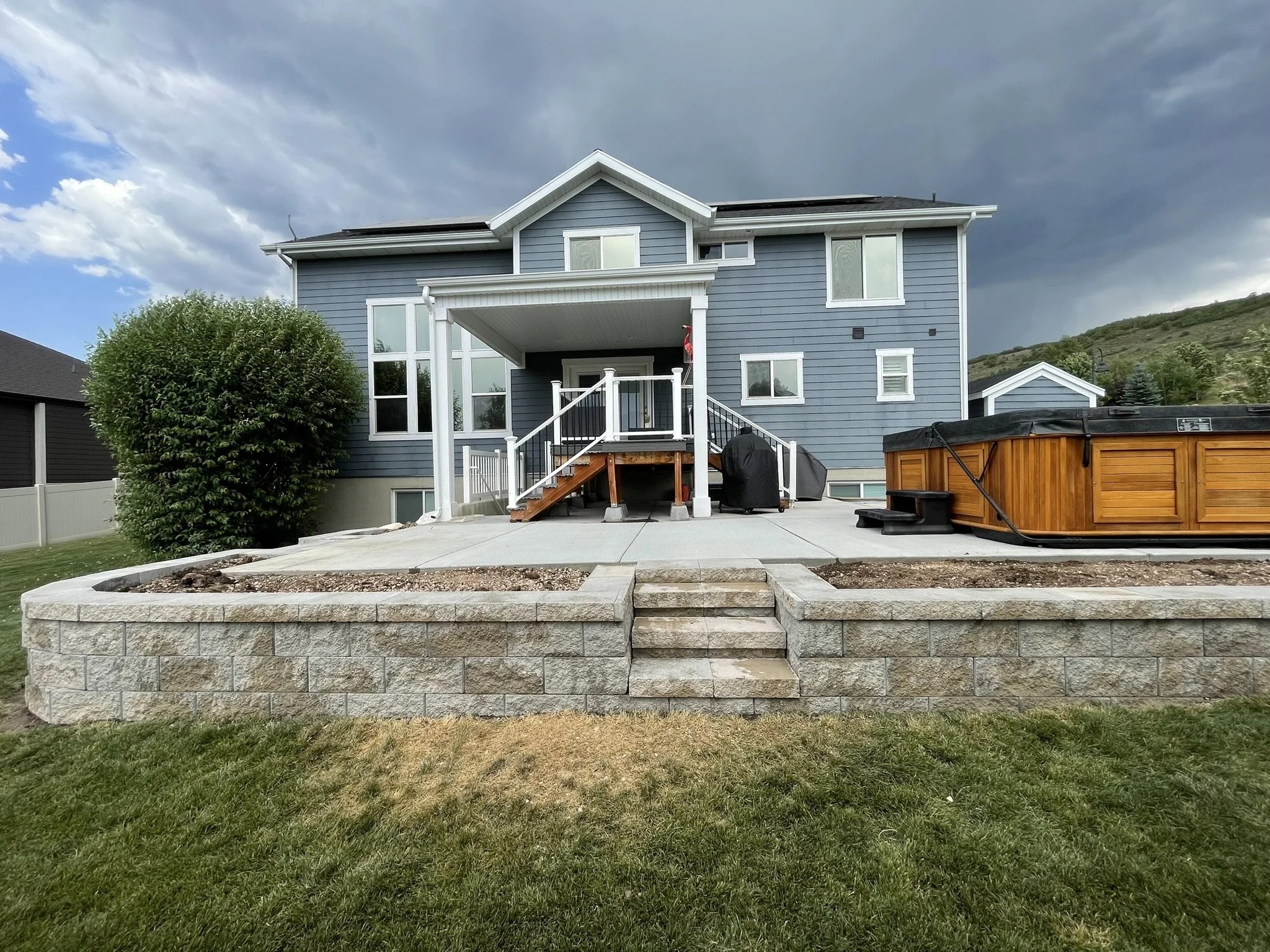Backyard of a blue two-story house with a patio, steps, hot tub, and landscaped garden area.