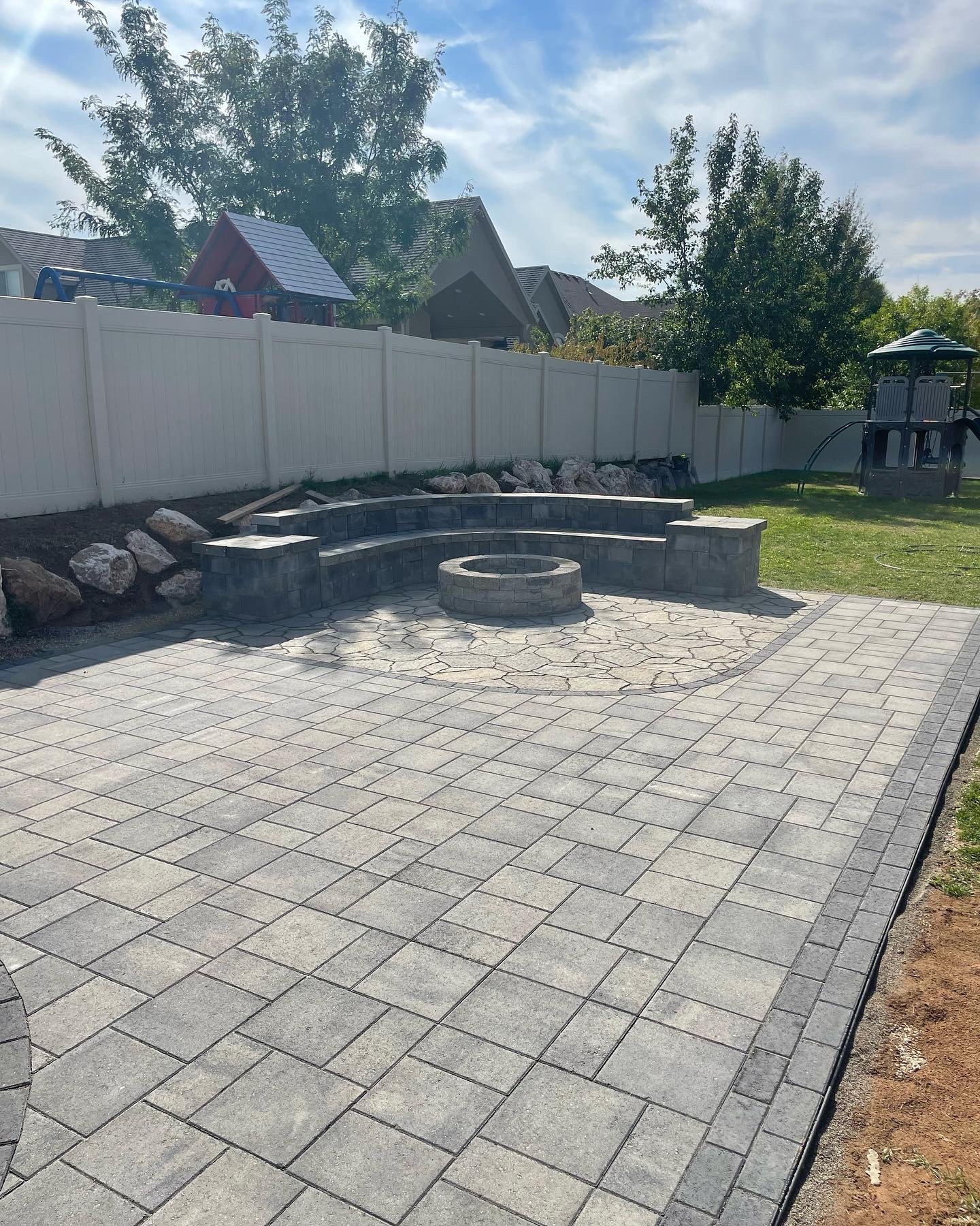 Backyard with newly paved stone patio, a circular fire pit, a curved stone bench, a white fence, a children's playhouse, trees, and a play structure with slide in the background.