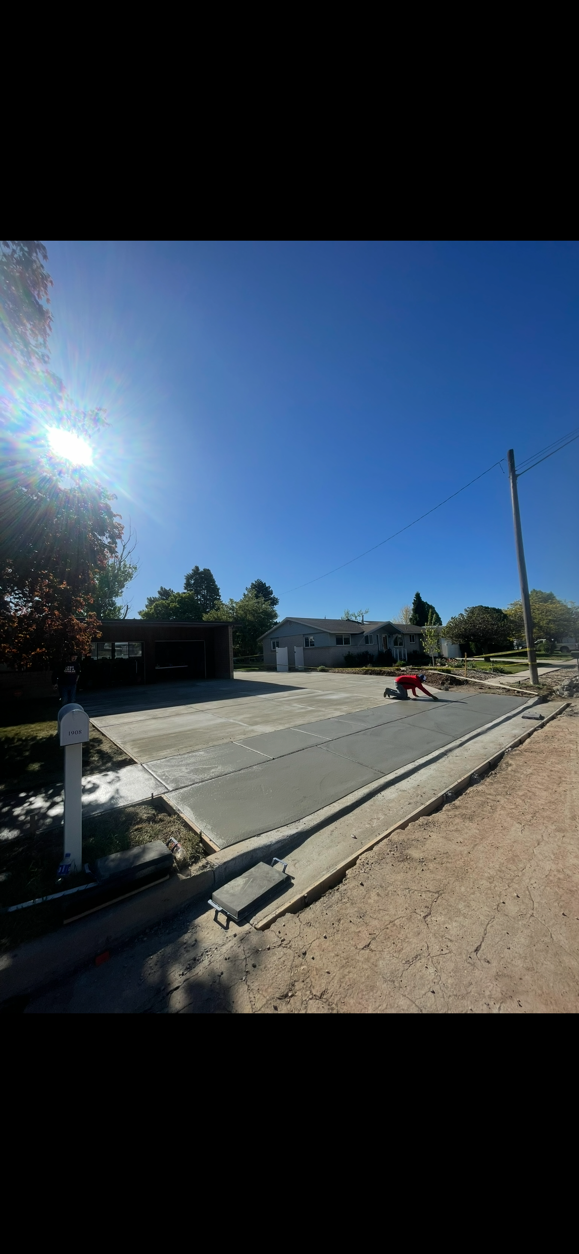 Concrete driveway being poured in a residential neighborhood on a sunny day, with houses, trees, and a utility pole in the background.