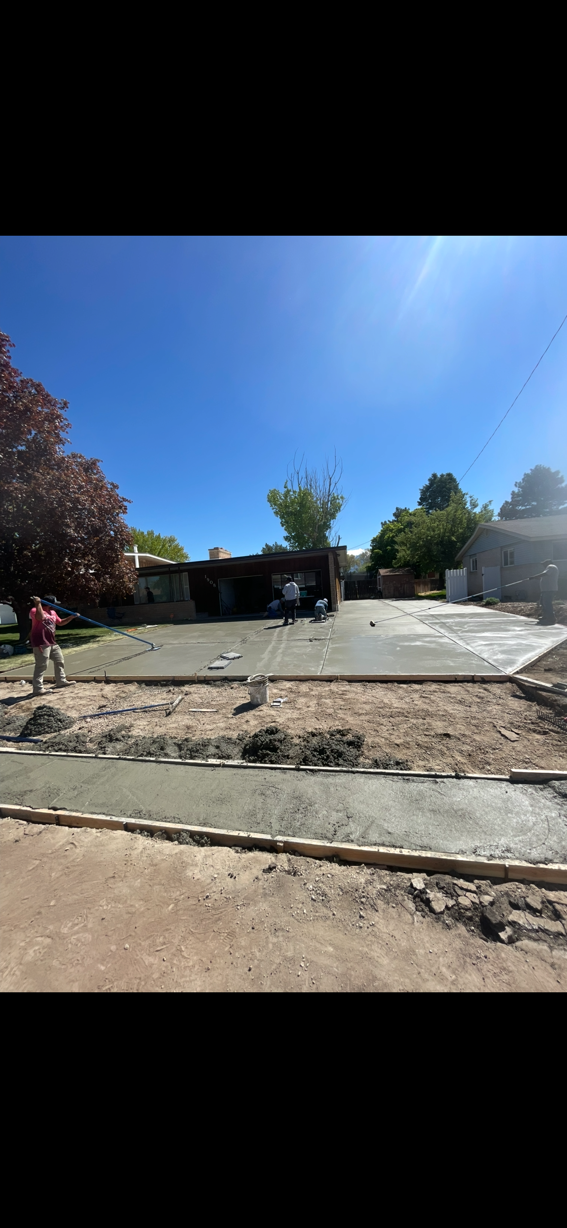 People pouring and smoothing concrete for a driveway or patio in a residential backyard on a sunny day.