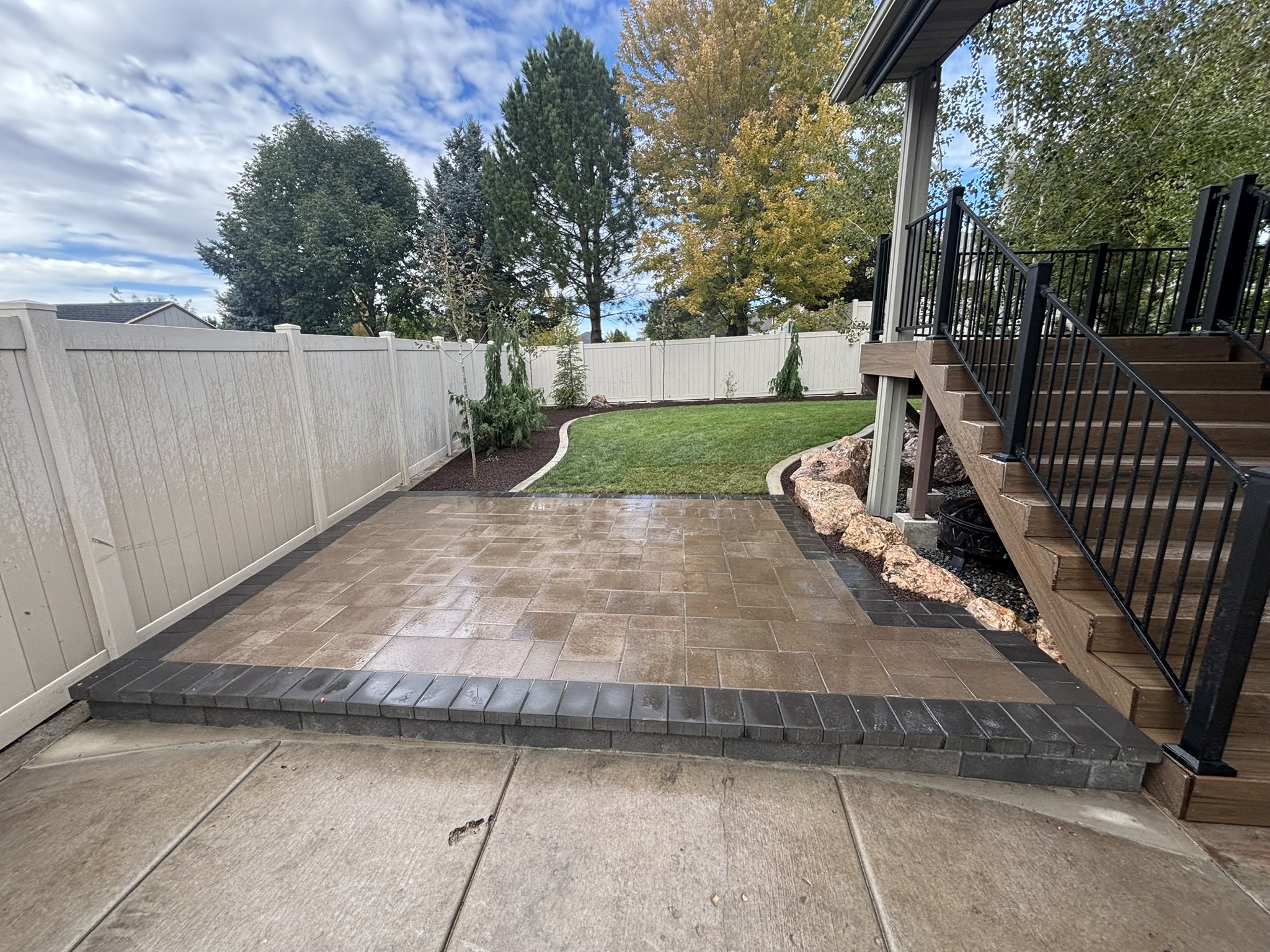 Backyard patio with tan pavers, black brick border, white vinyl fence, landscaped yard with small trees, grass, and rocks, wooden stairs with black railing leading to the house, and a cloudy sky.