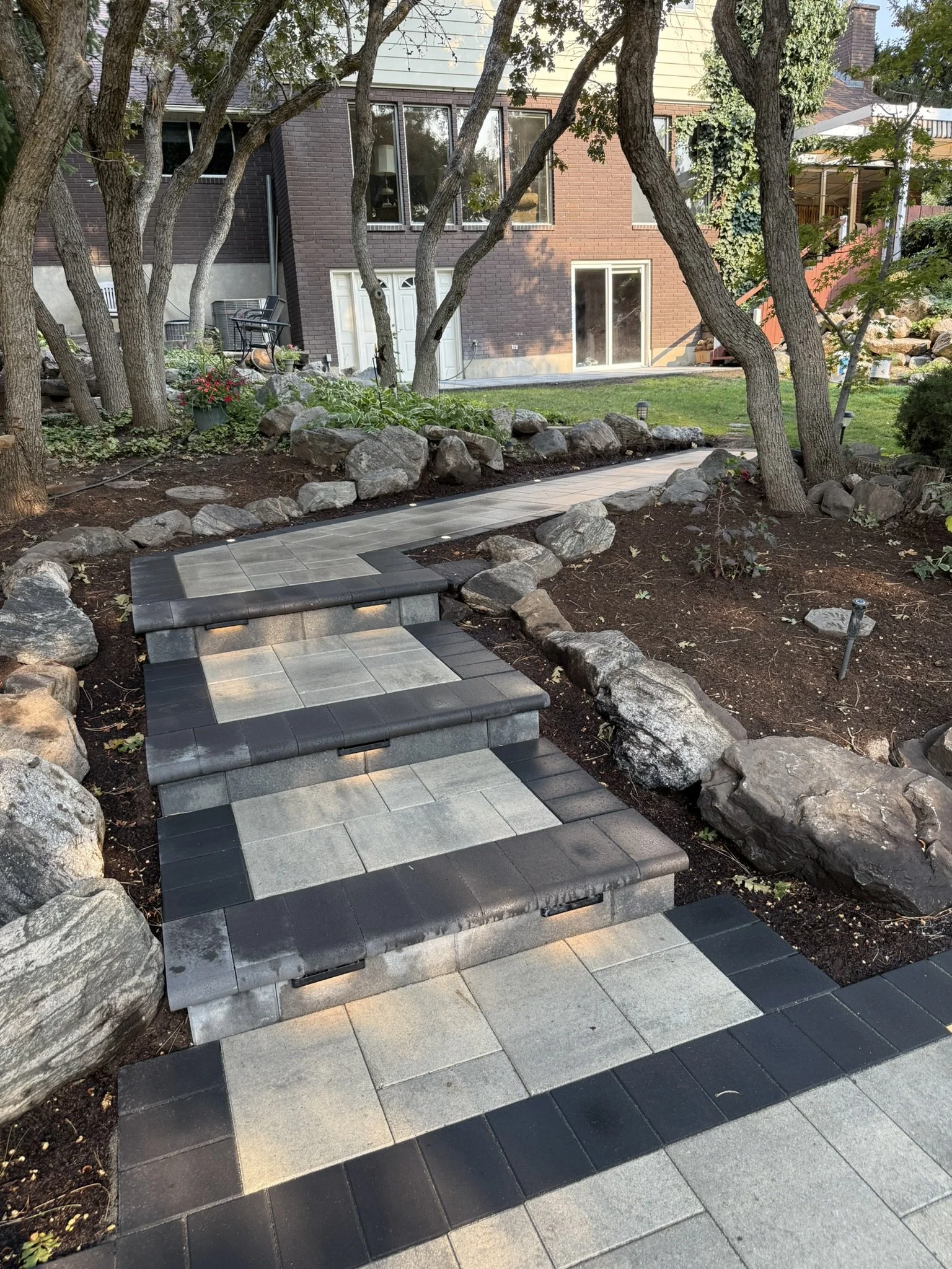A stone pathway with steps leading up a garden, bordered by rocks, with trees and a house in the background.