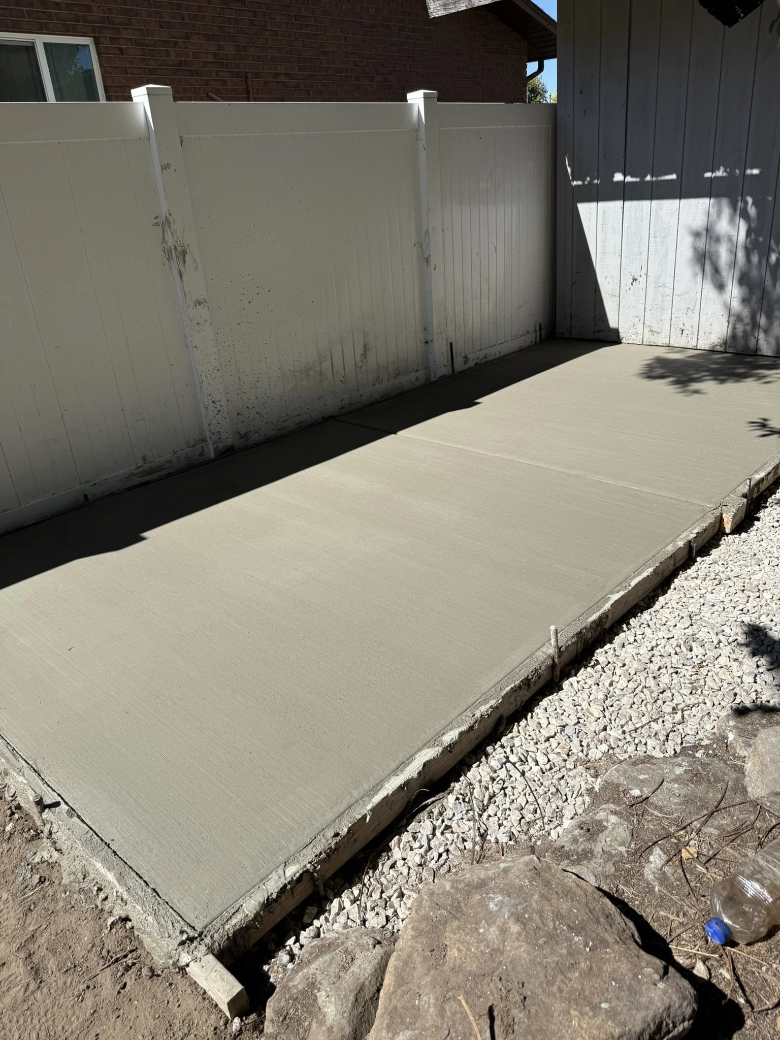 Recently poured concrete patio next to a white wooden fence with some dirt and rocks along the edges.