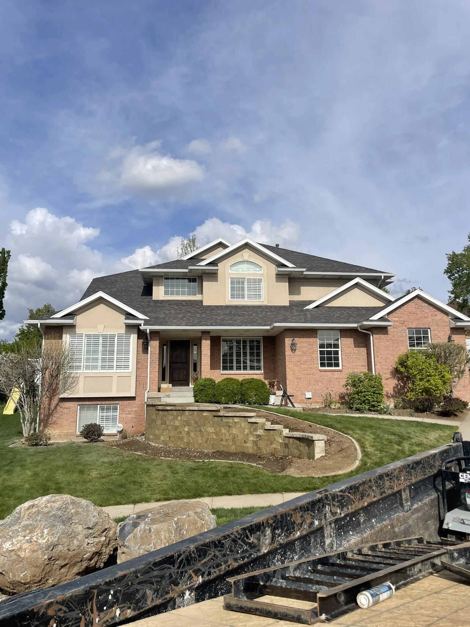 Large two-story brick house with front porch, steps, and a curved retaining wall, surrounded by grass and shrubbery, under a partly cloudy sky.