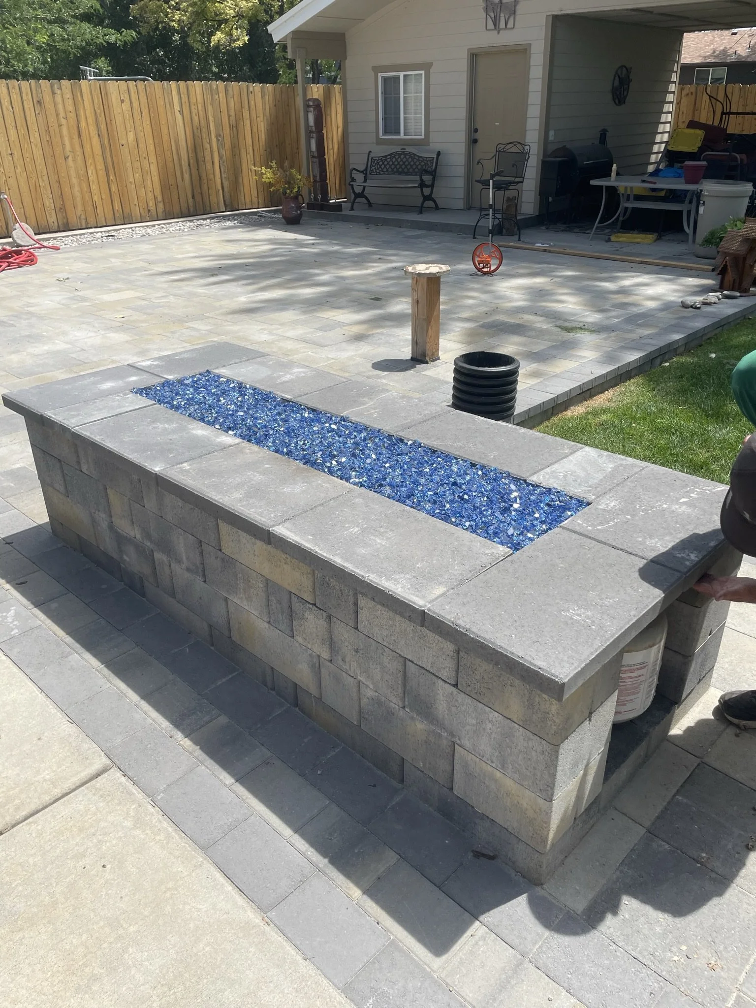 A newly built outdoor fire pit with a rectangular blue glass inset, constructed from gray stone blocks, in a backyard patio area under construction.
