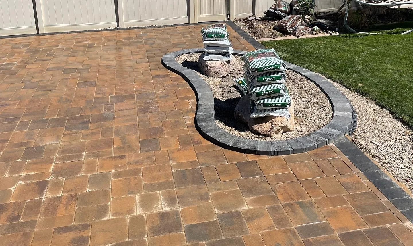 A newly paved outdoor patio with interlocking brick tiles, a curved border, and a small landscaped area with rocks and bags of soil or fertilizer on top.