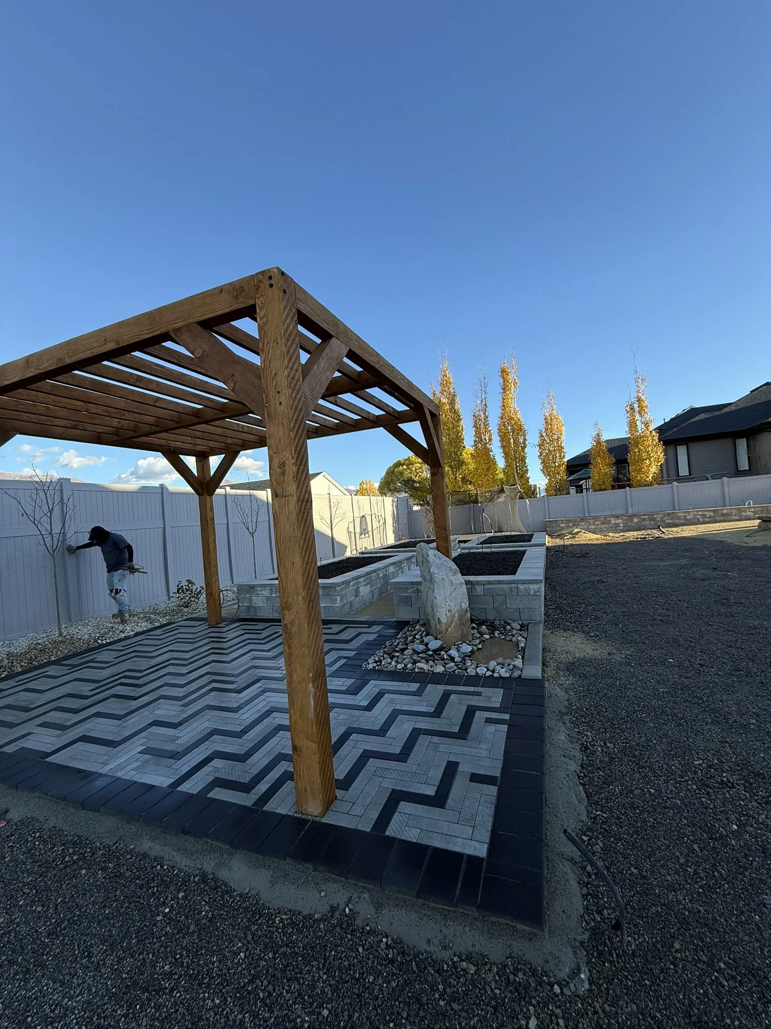 A backyard with a wooden pergola, patterned gray and black pavers, a white stone feature, and a light gray fence, with a person planting a tree in the background.